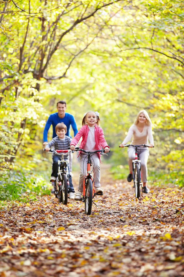 Fall Family 桌布 壁紙 — 一個幸福的四口之家在鋪滿落葉的森林小徑上騎腳踏車，沐浴在金色的秋日陽光中。