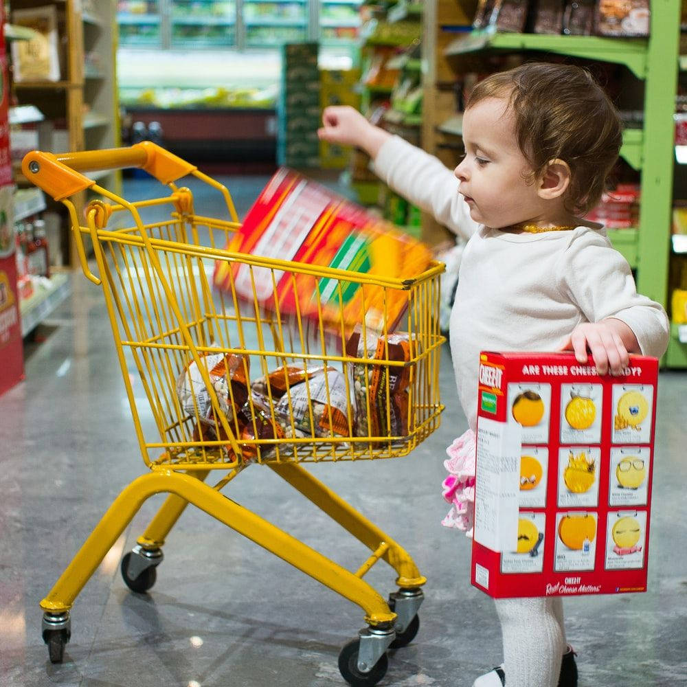 金錢 桌布 壁紙 — 一個幼兒在光線明亮的雜貨店通道中，推著一輛裝滿零食的黃色購物車，同時拿著一盒起司午間小食。
