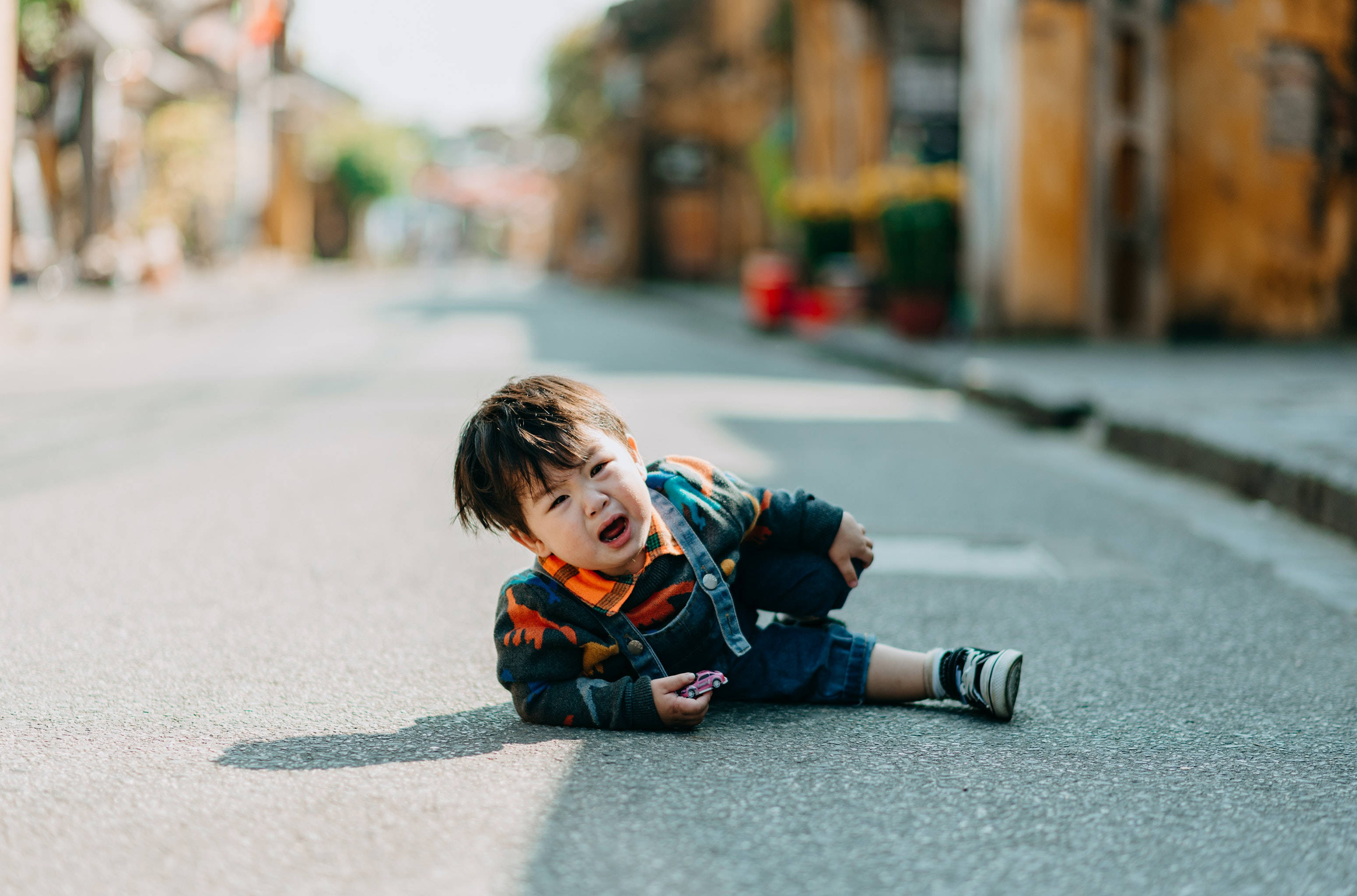 crying sad boy 桌布 壁紙 — 一個幼兒坐在陽光普照的街道上哭泣，背景是模糊的暖色調建築物。