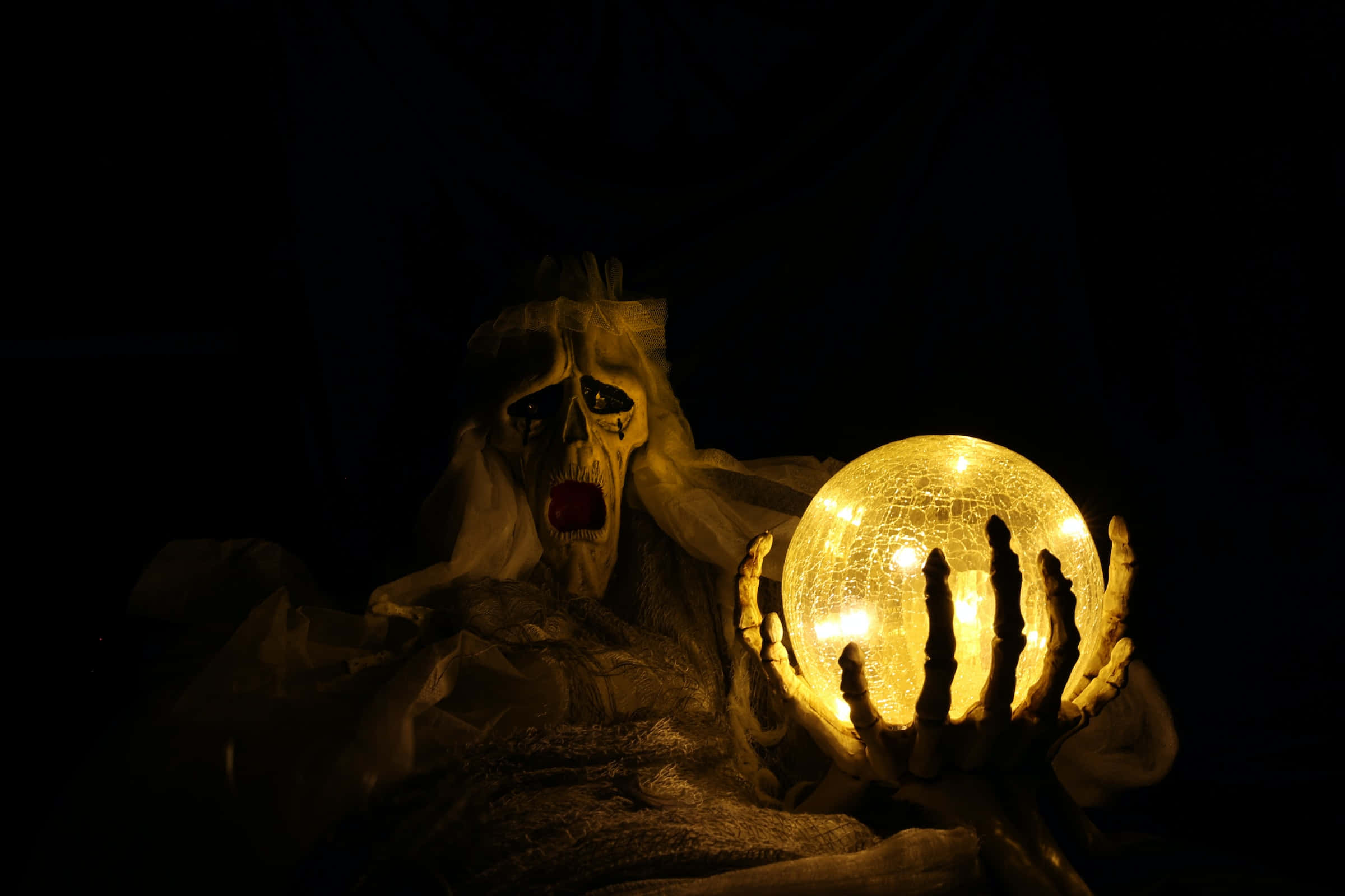 Halloween Desktop 桌布 壁紙 — 一個幽靈般的人物，有著骷髏臉和破爛的面紗，在黑暗的環境中拿著一個發光且破裂的水晶球。