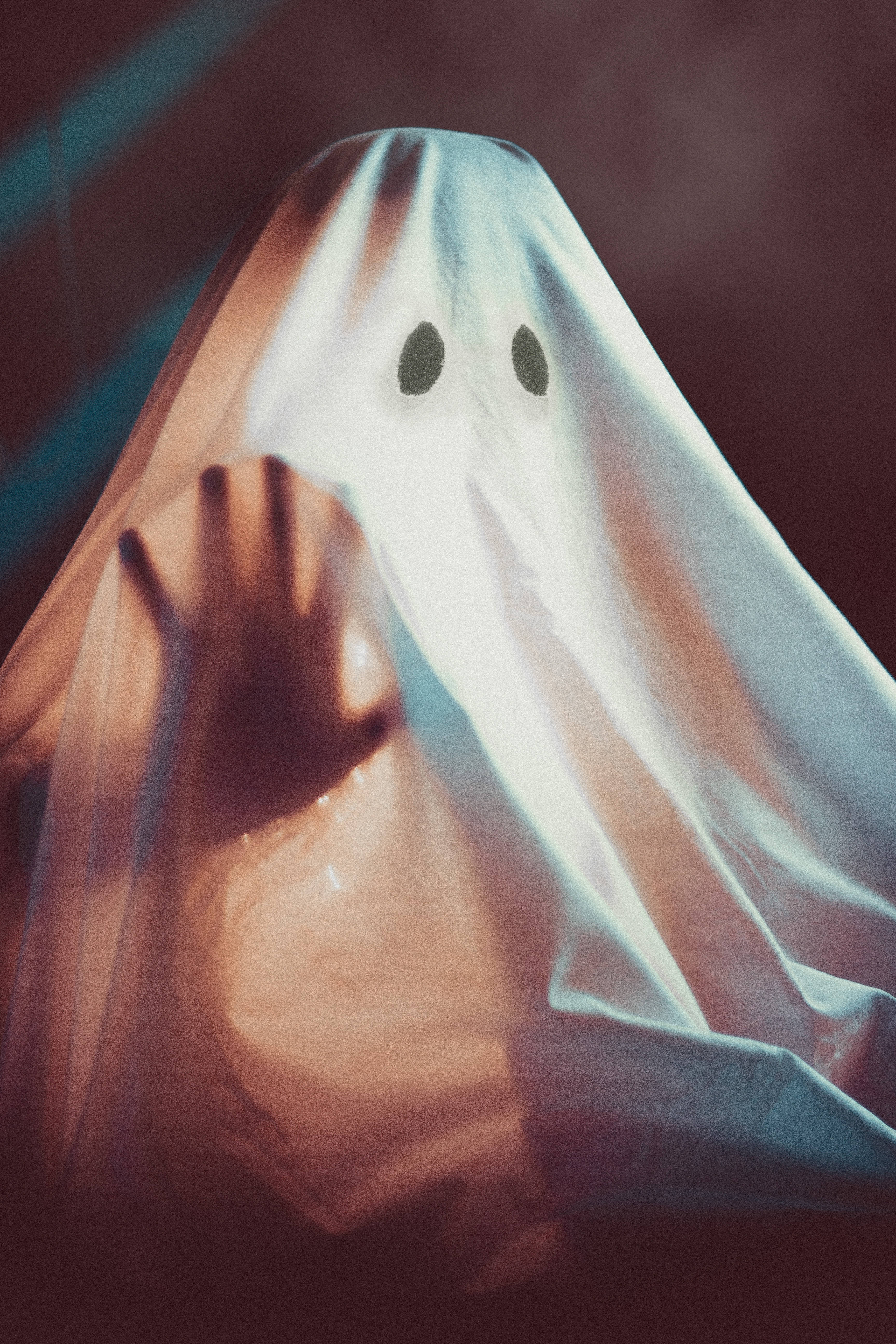Halloween Phone 桌布 壁紙 — 一個幽靈般的身影，披著帶黑色眼孔的白布，在昏暗、充滿氣氛的環境中舉起一隻手。