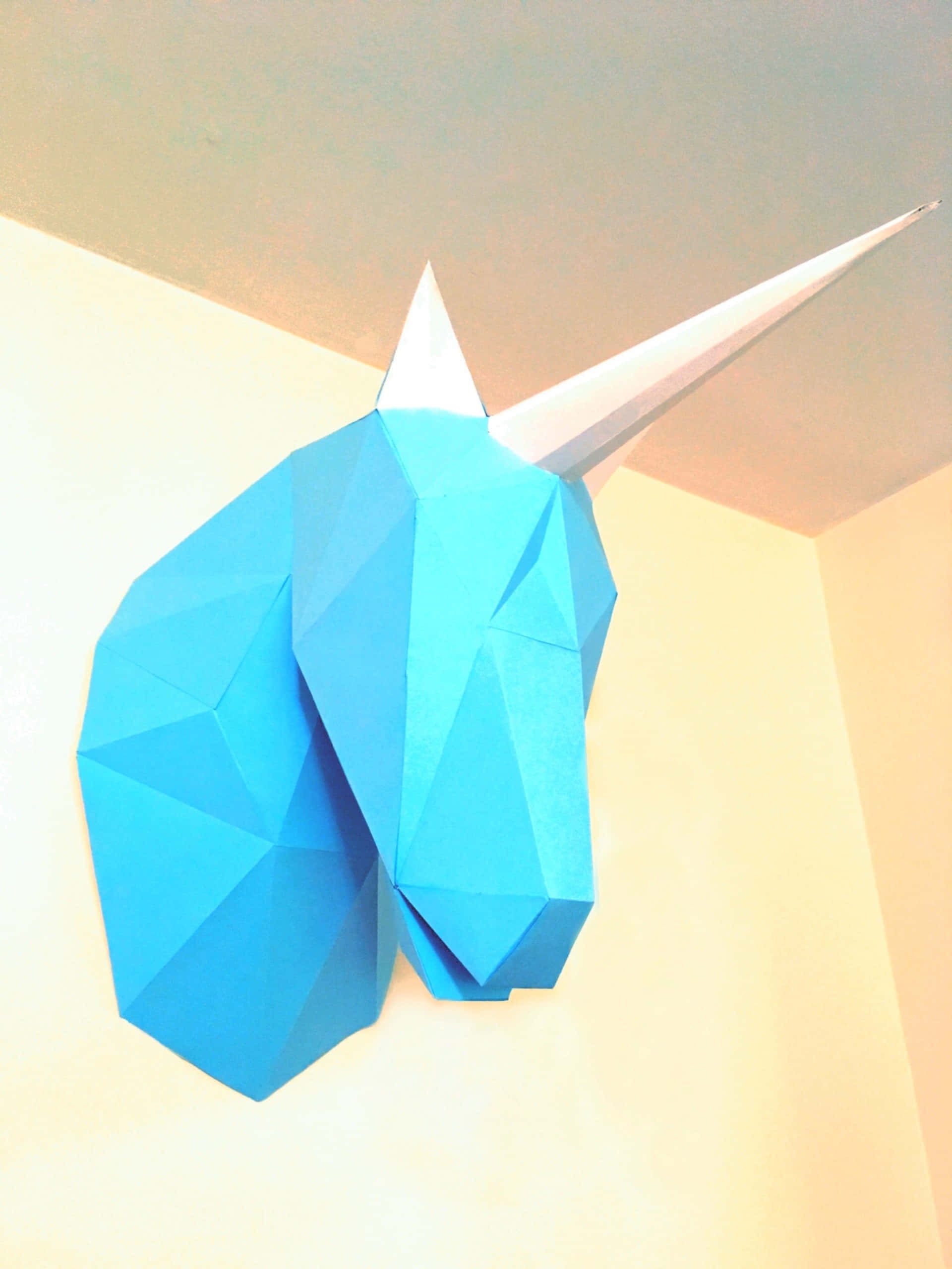 3d Unicorn 桌布 壁紙 — 一個幾何形狀的藍色紙獨角獸頭部，帶有白色角，安裝在溫暖環境照明的牆上。