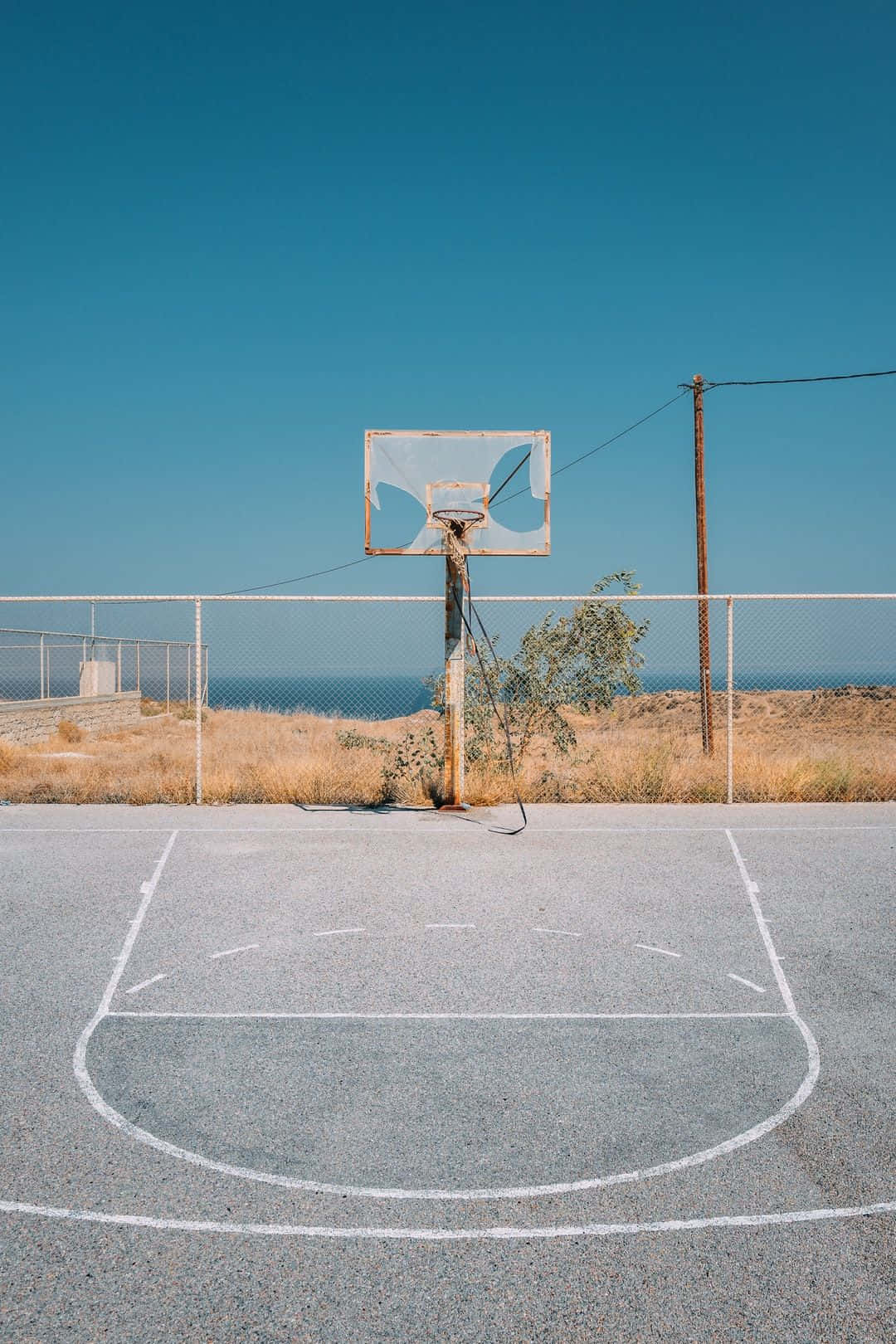 Basketball Court 桌布 壁紙 — 一個廢棄的籃球場，帶有褪色的線條和一個破損的籃框，矗立在晴朗的藍天下，俯瞰海洋。