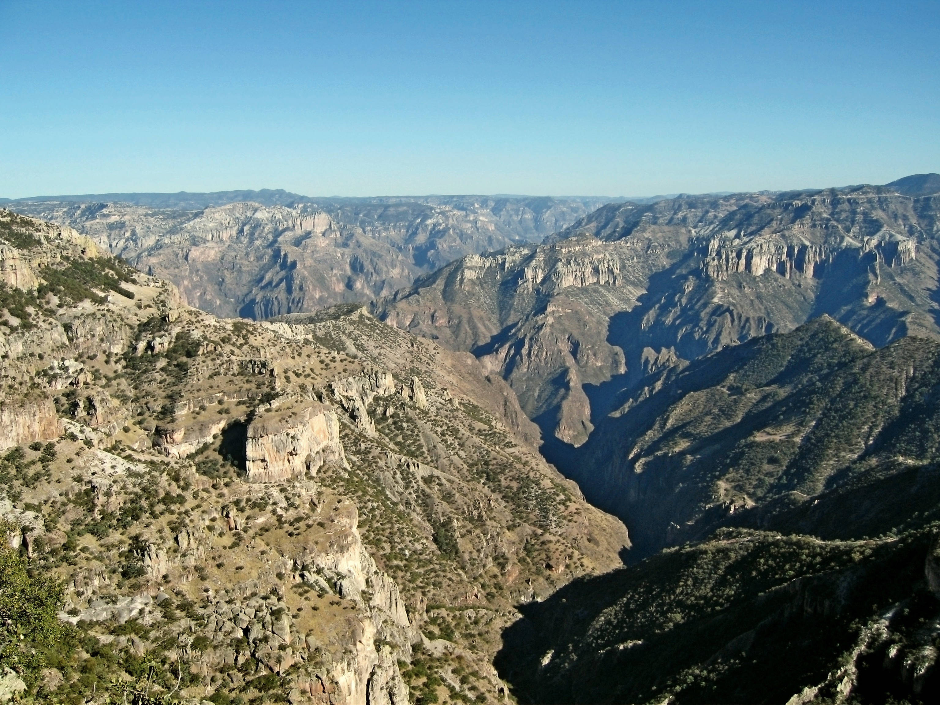 Canyon 桌布 壁紙 — 一個廣闊、陽光普照的峽谷，擁有崎嶇的懸崖和層狀岩層，在清澈的藍天下延伸。