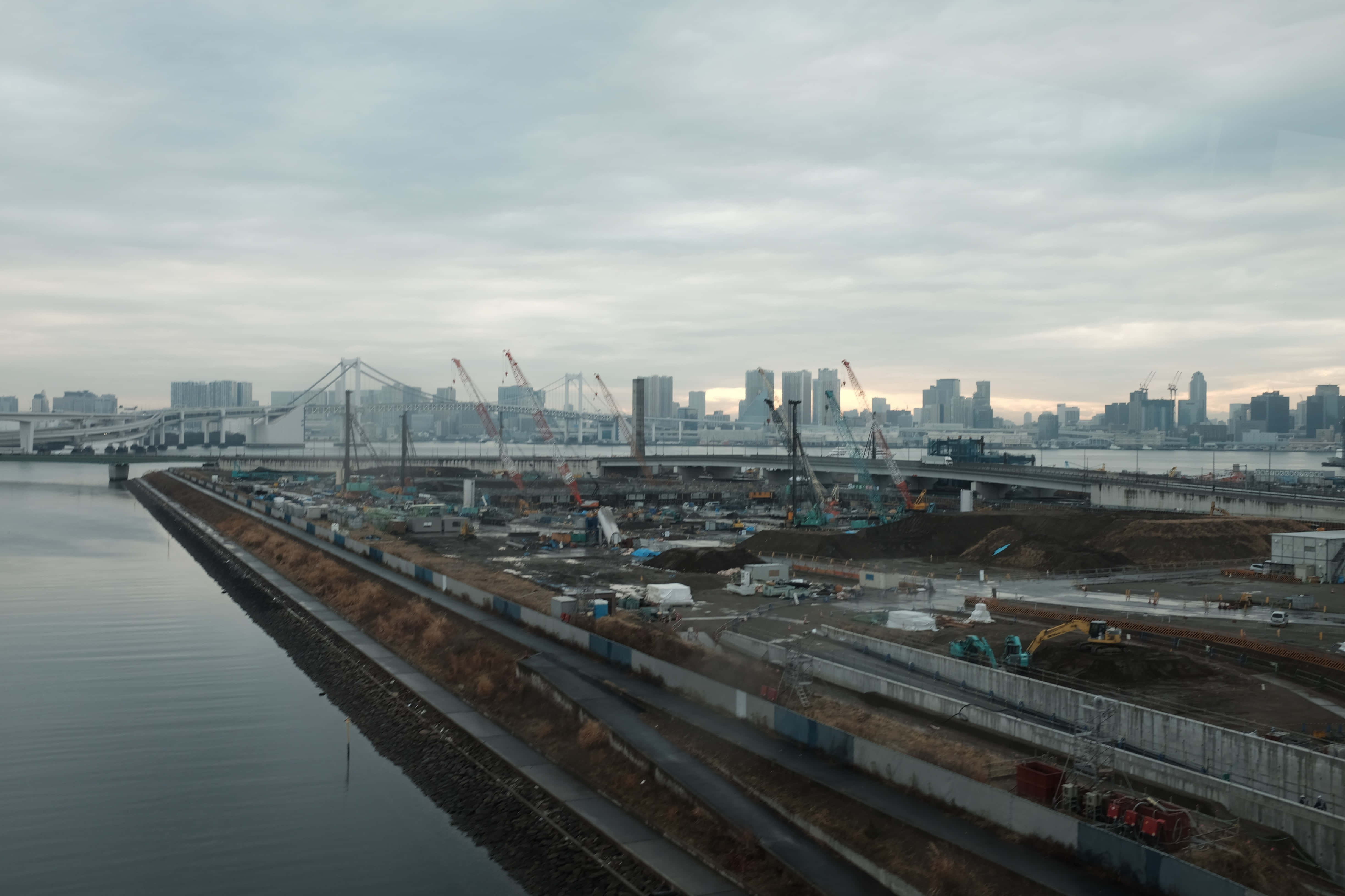 Infrastructure 桌布 壁紙 — 一個廣闊的城市建築工地沿著平靜的河流延伸，在陰天之下，有起重機和遠處的城市摩天大樓。