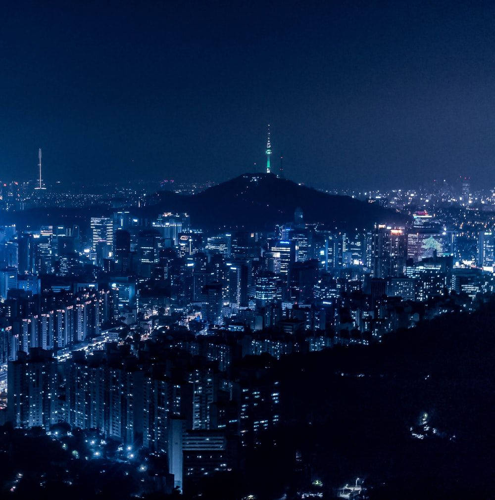 korean 桌布 壁紙 — 一個廣闊的夜景城市景觀，閃爍的摩天大樓和一座山頂上有發光塔樓，在深藍色的天空下。