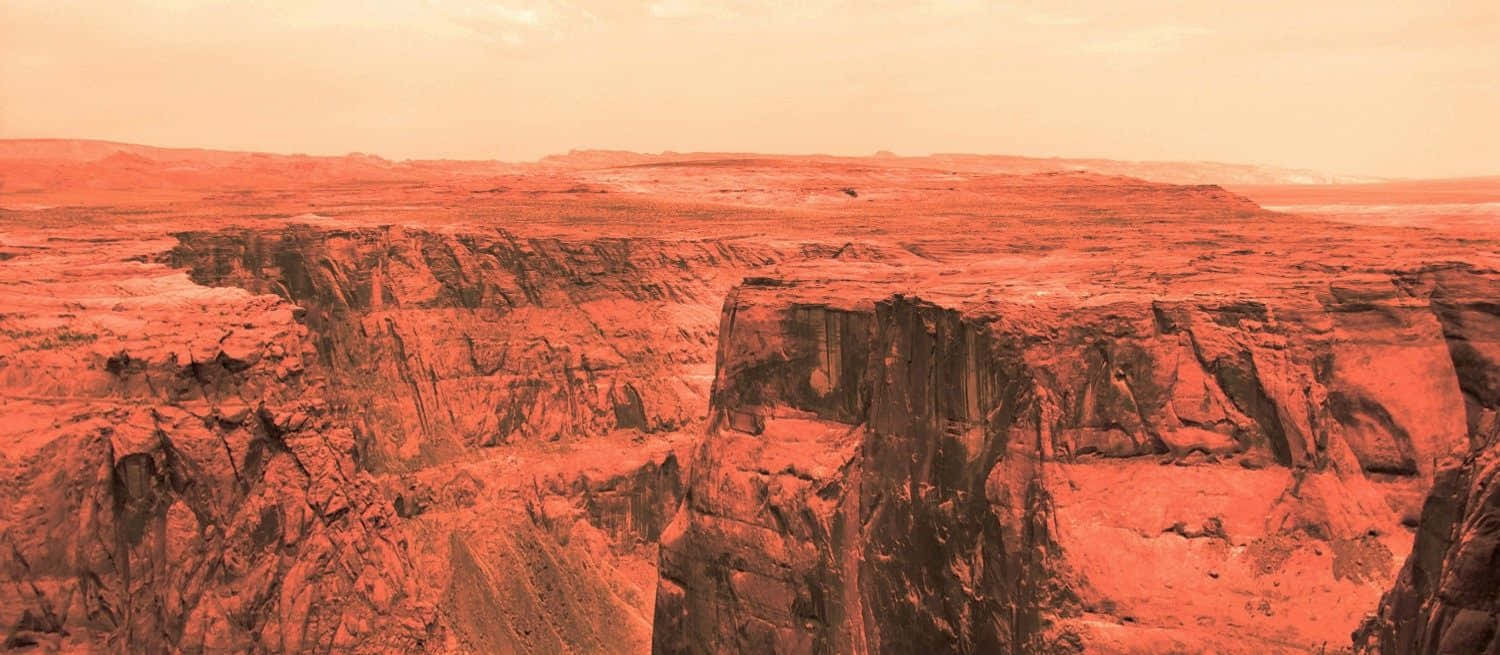 Mars Landscape 桌布 壁紙 — 一個廣闊的峽谷，陡峭的紅色岩壁在朦朧的橙色天空下延伸。