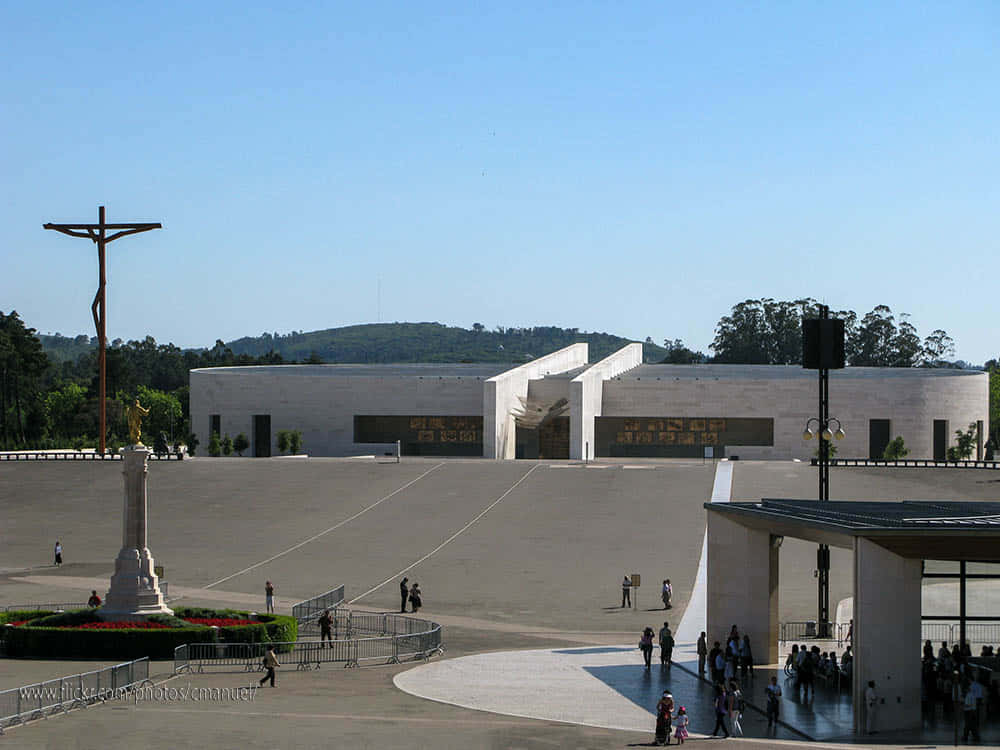 Portugal 桌布 壁紙 — 一個廣闊的廣場，聳立著十字架和現代白色教堂，在晴朗的藍天下，遠處有綠色的山丘。