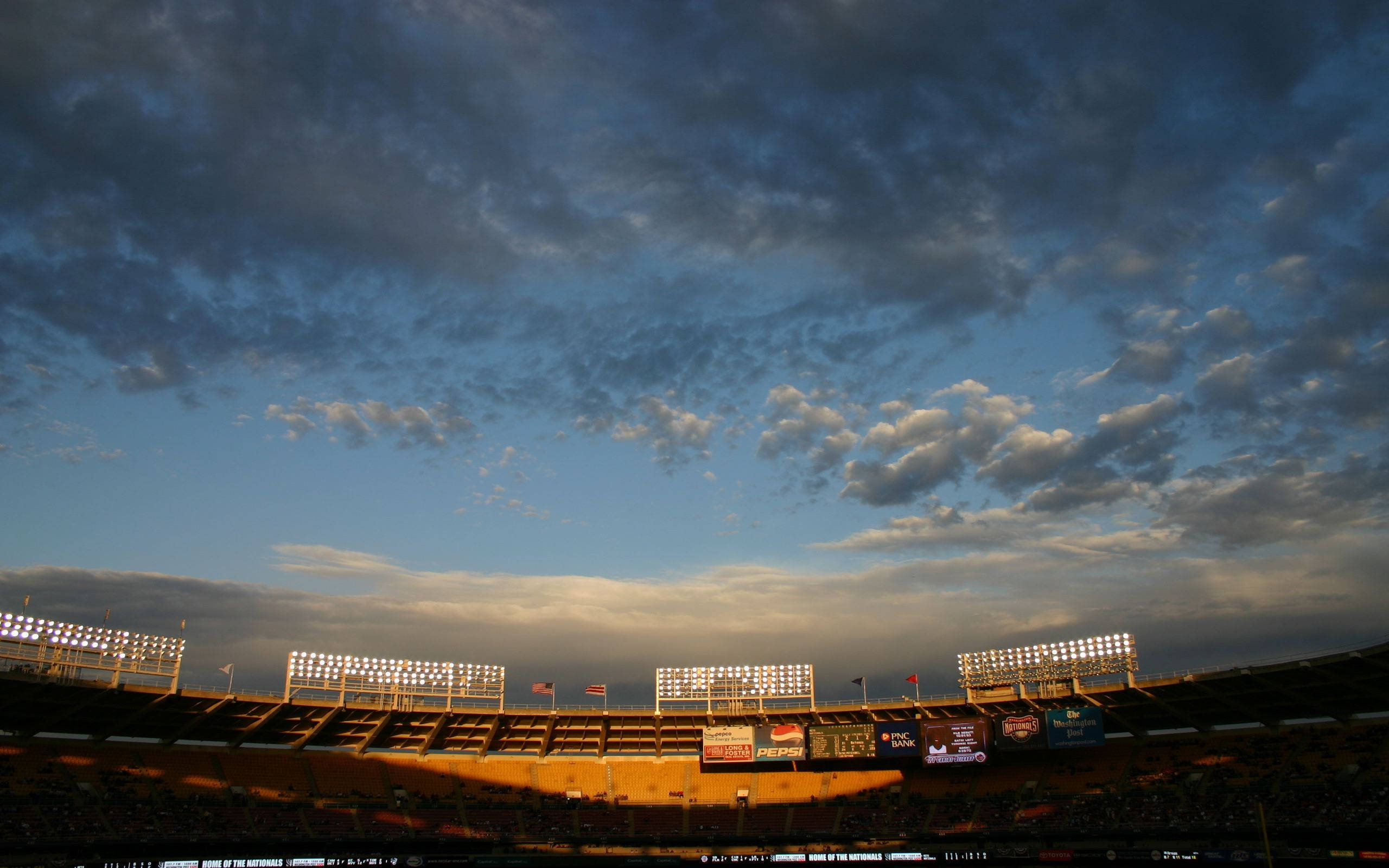 Nationals 桌布 壁紙 — 一個廣闊的棒球場沐浴在金色的日落光線中，上方有戲劇性的雲層，發光的泛光燈照亮了空蕩蕩的看台。