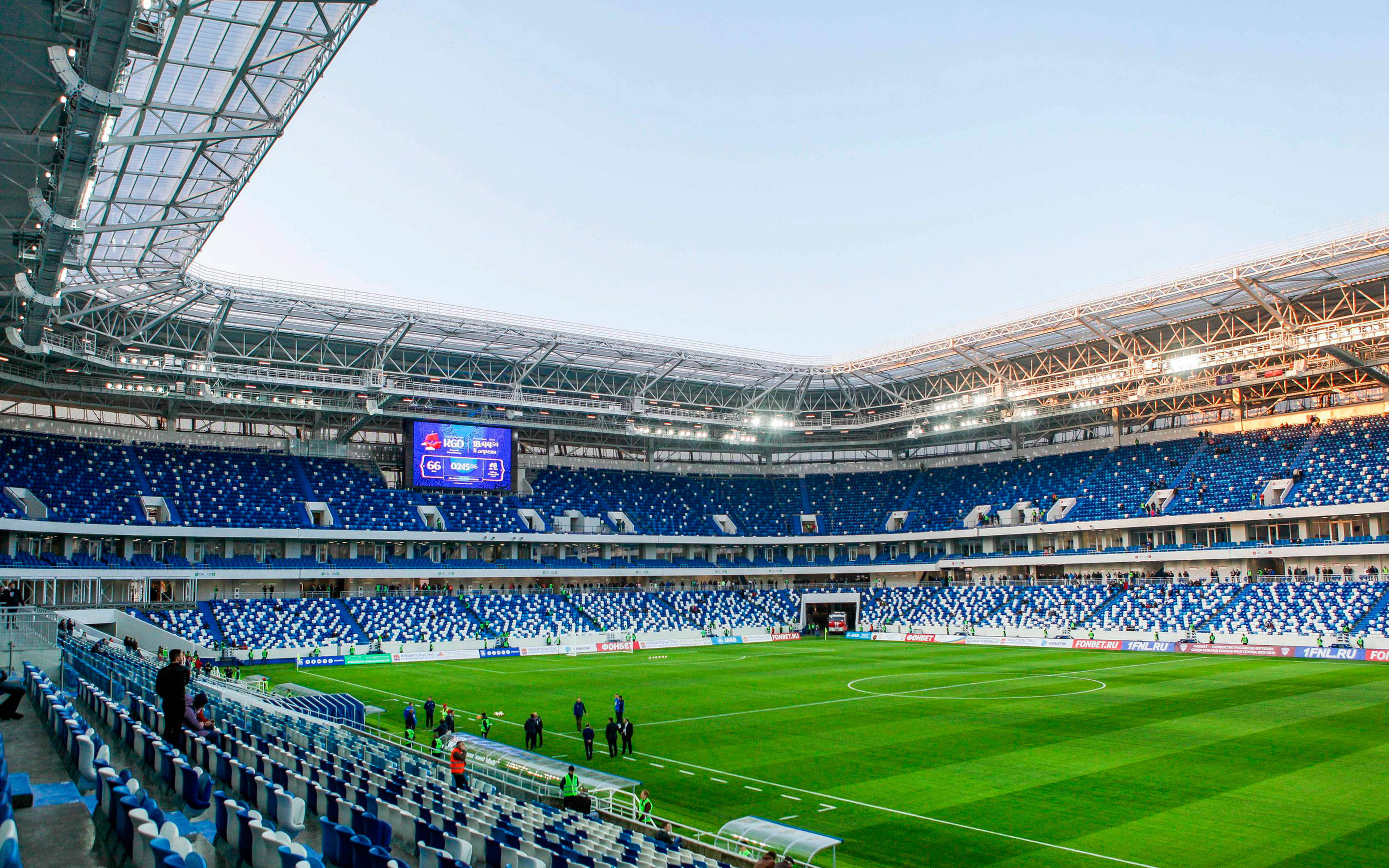 Stadium 桌布 壁紙 — 一個廣闊的現代足球場，設有藍色座位和充滿活力的綠色草坪，在晴朗的天空下。