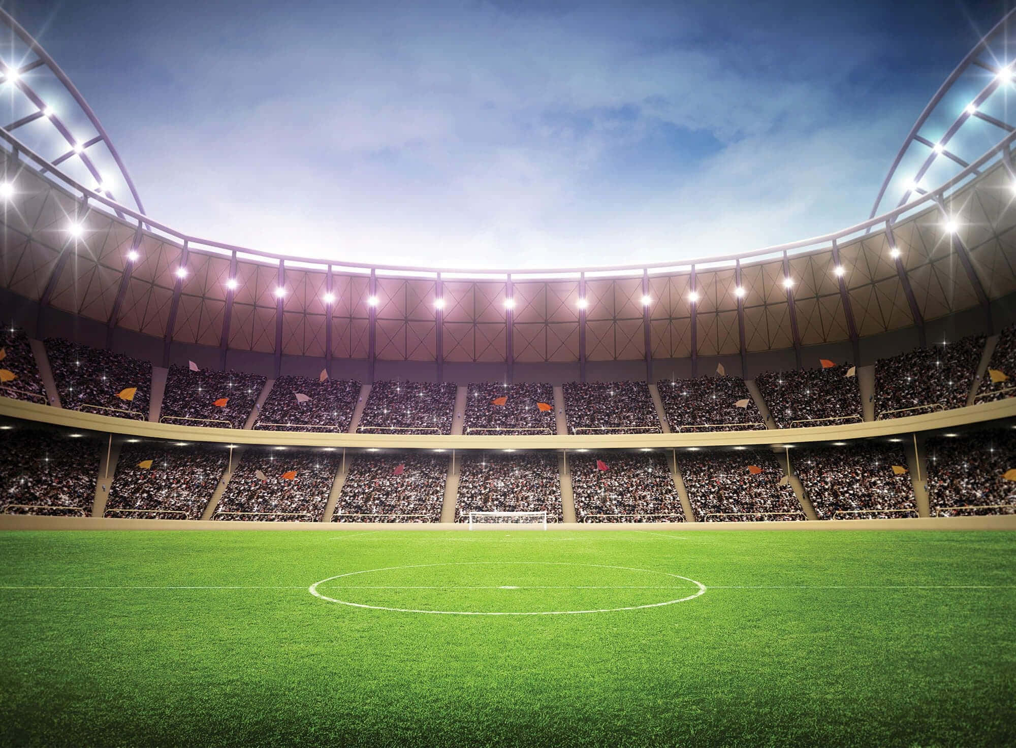 Stadium 桌布 壁紙 — 一個廣闊的綠色足球場，在明亮的體育場燈光下，看台座無虛席，上方是清澈的藍天。