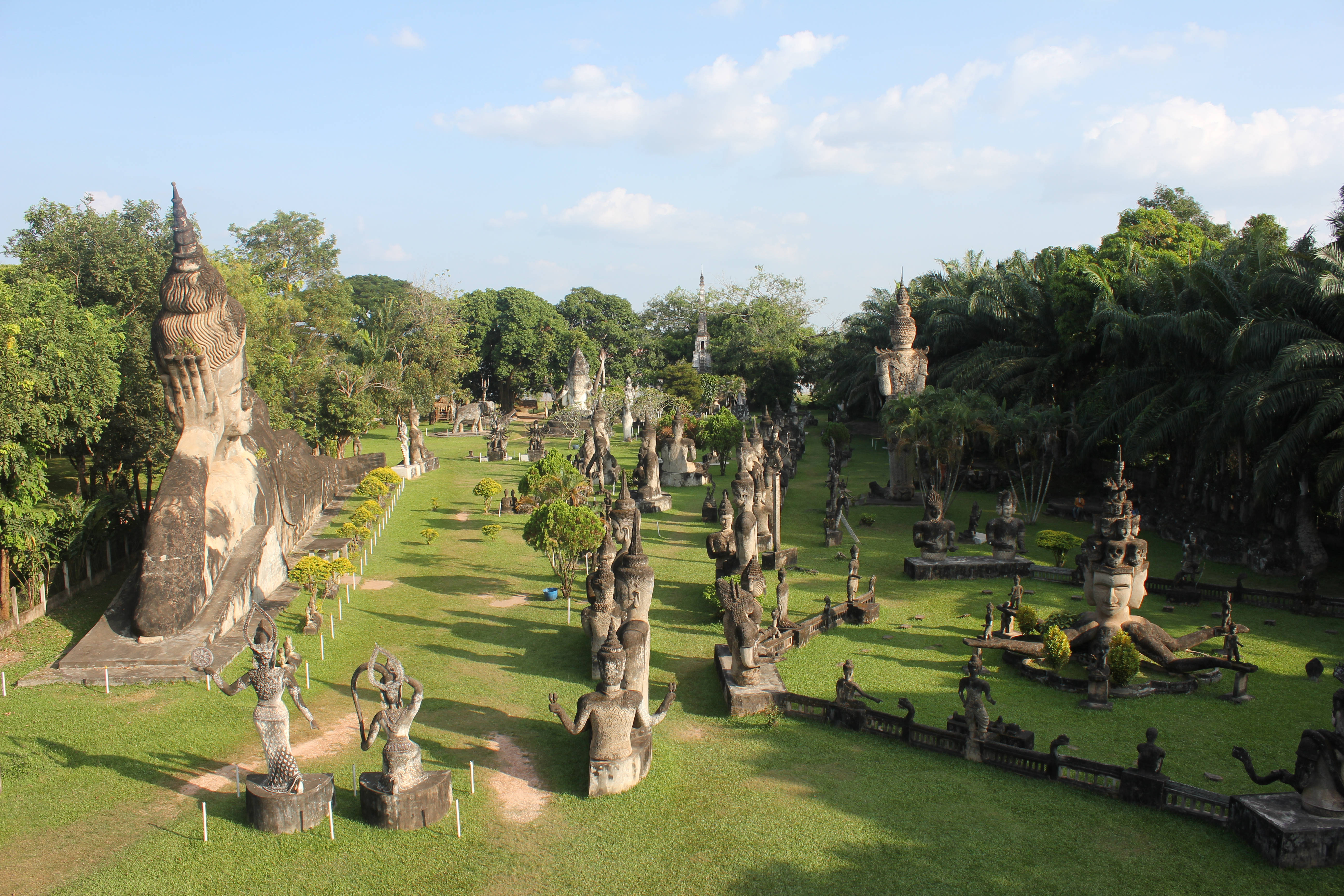 Laos 桌布 壁紙 — 一個廣闊的花園，充滿了古老的佛像和神話人物的石像，在明亮的藍天和零星雲朵下。