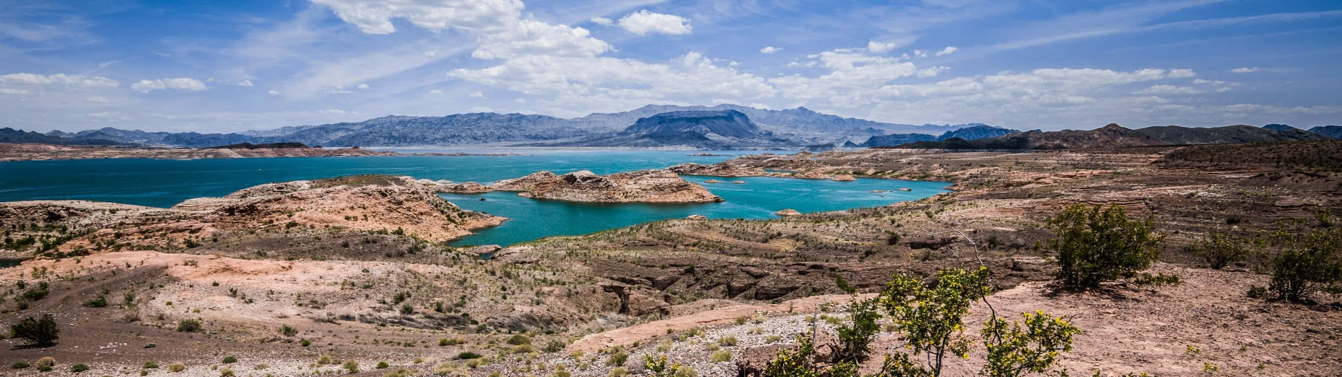 Lake 桌布 壁紙 — 一個廣闊的藍綠色湖泊，坐落在乾旱的沙漠地形中，遠處有山脈，在明亮的藍天下有散落的雲朵。