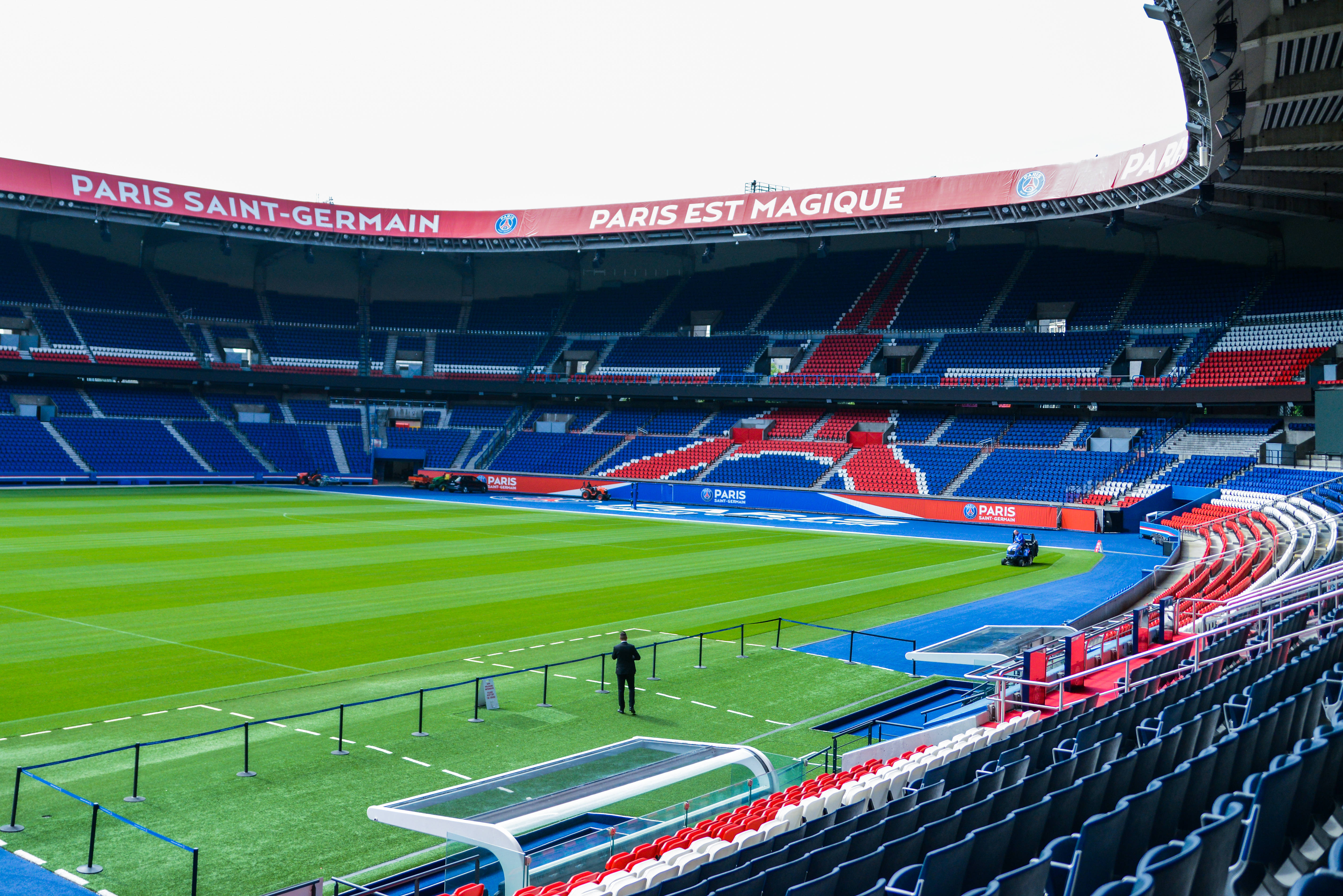 梅西 PSG 桌布 壁紙 — 一個廣闊空曠的巴黎聖日耳曼球場，擁有充滿活力的綠色草坪和藍紅色的座位，天空多雲。