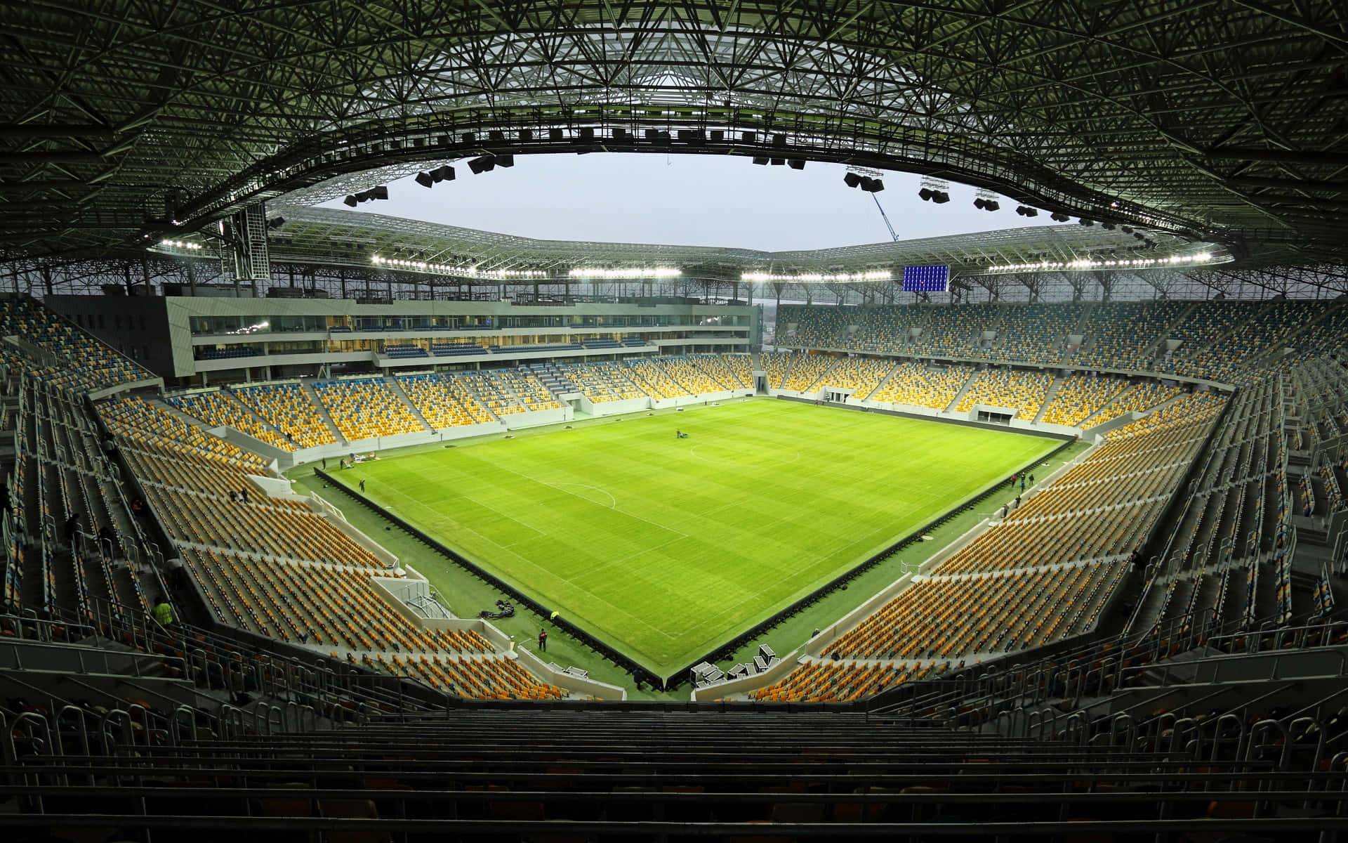 Stadium 桌布 壁紙 — 一個廣闊空蕩的足球場，在鋼鐵屋頂下，黃昏時分有充滿活力的綠色草坪和黃色座位。