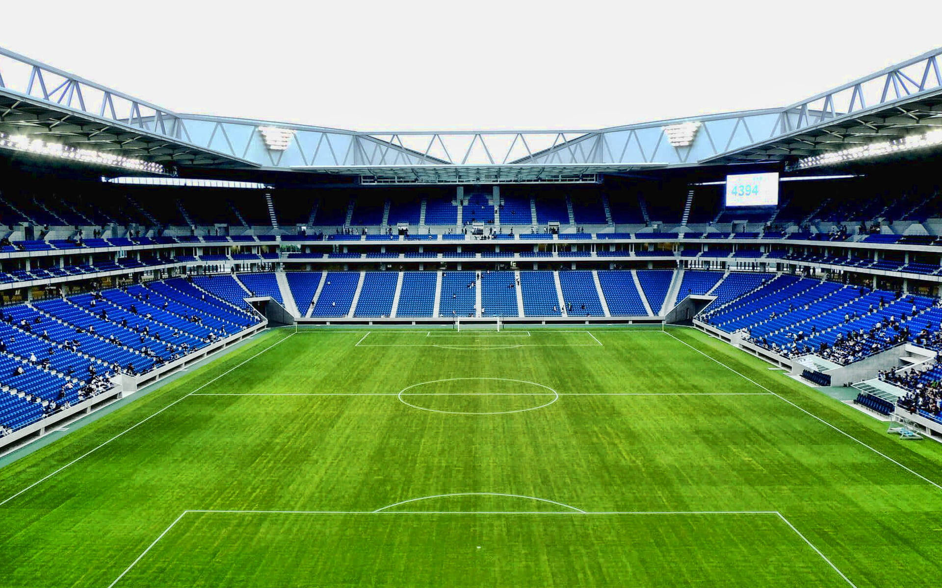 Stadium 桌布 壁紙 — 一個廣闊空蕩的足球場，擁有鮮綠色草地和藍色座位排，在明亮的天空下。