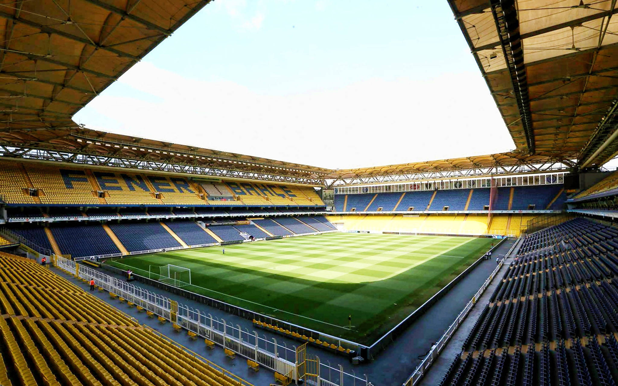 Stadium 桌布 壁紙 — 一個廣闊空蕩的足球場，黃色和藍色座位，在明亮的白天天空下。