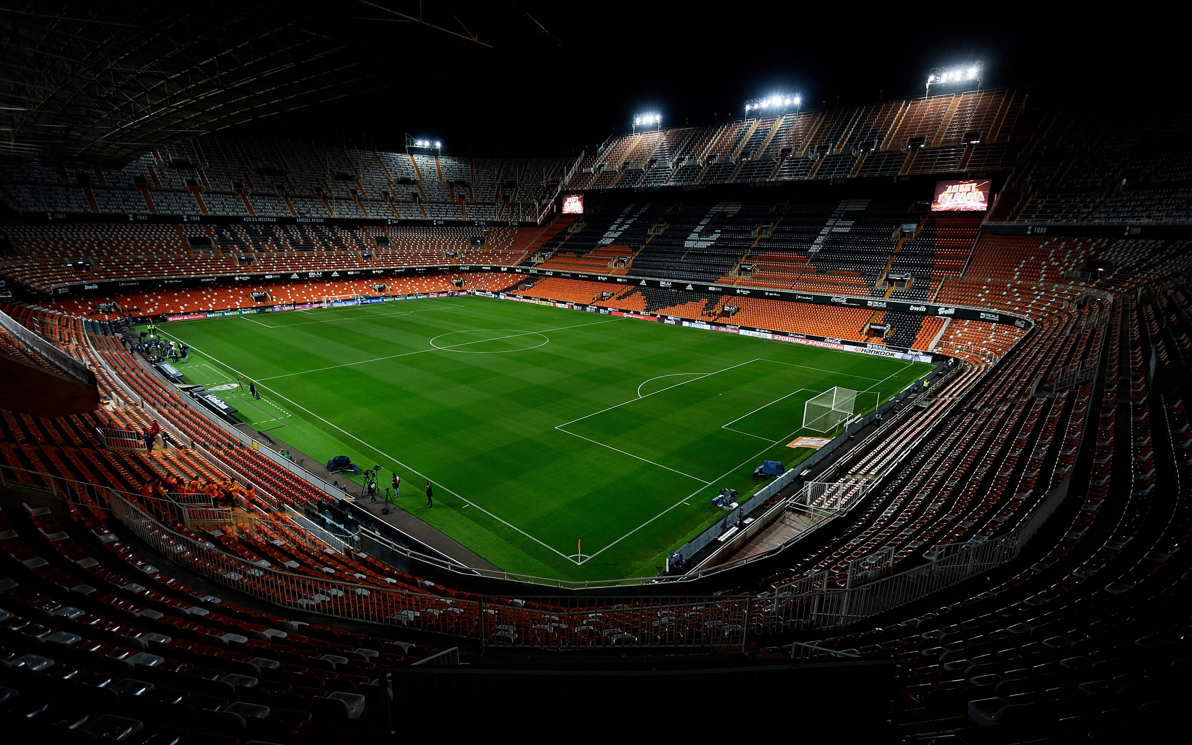 Stadium 桌布 壁紙 — 一個廣闊空蕩的足球場在夜晚被明亮的泛光燈照亮，在充滿活力的綠色草坪和陶土色座位上投下銳利的陰影。