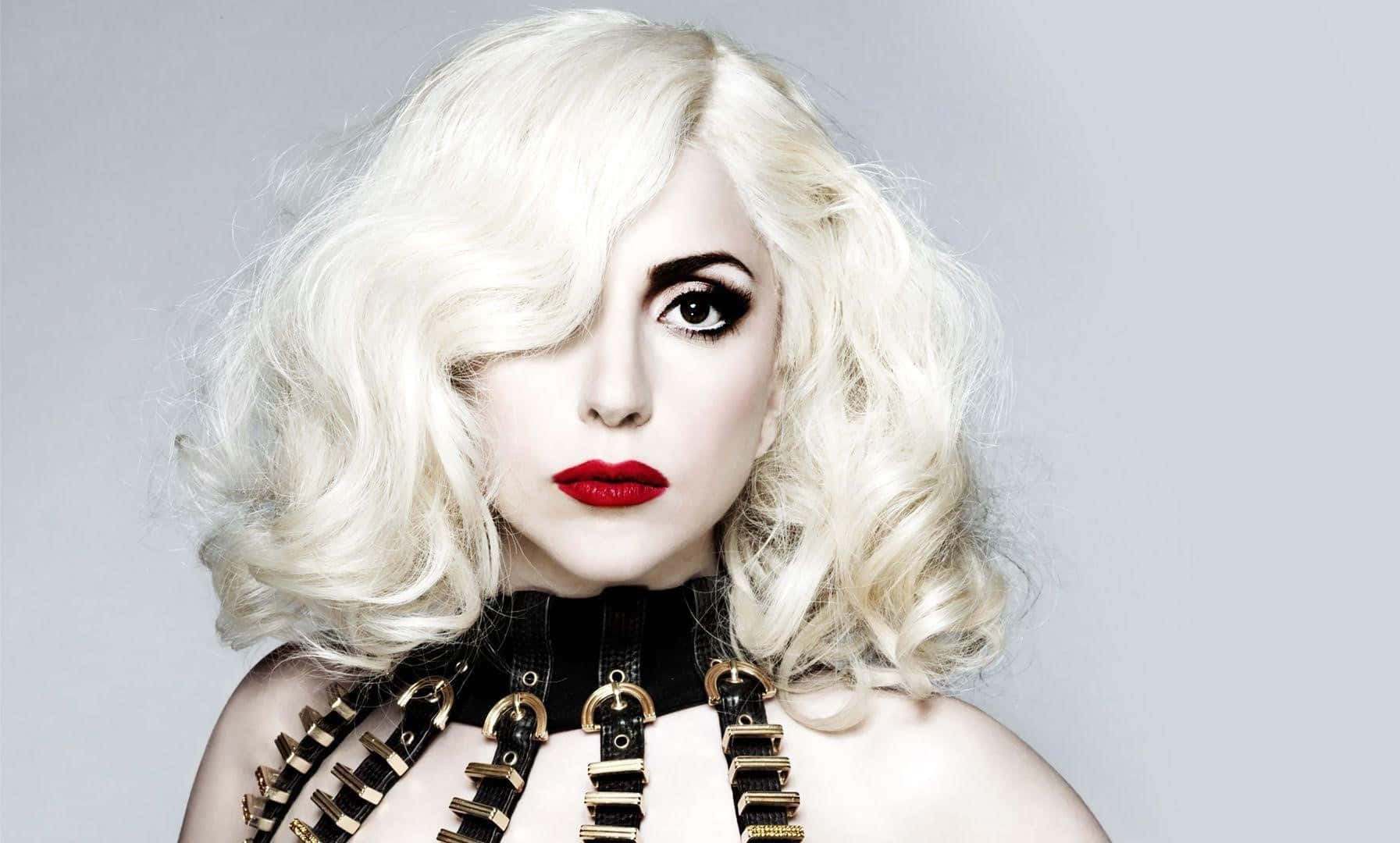 女神卡卡 桌布 壁紙 — 一個引人注目的女性肖像，擁有鉑金色捲髮、大膽的紅唇和黑色皮革背帶，背景是純灰色。