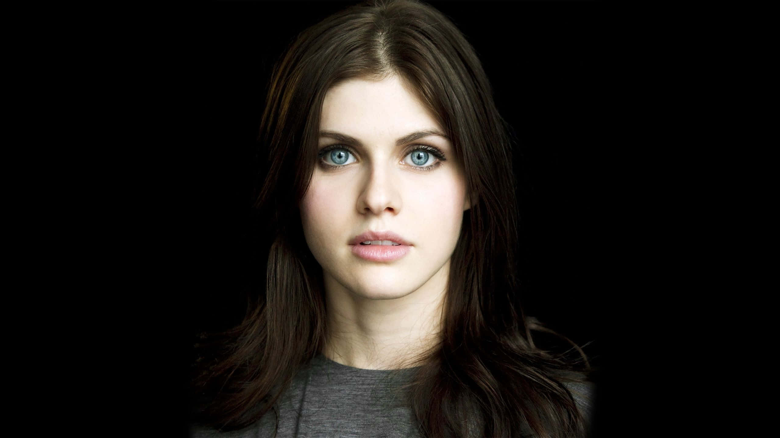 Alexandra Daddario 桌布 壁紙 — 一個引人注目的女性肖像，擁有鮮明的藍色眼睛和深色頭髮，背景為純黑色。