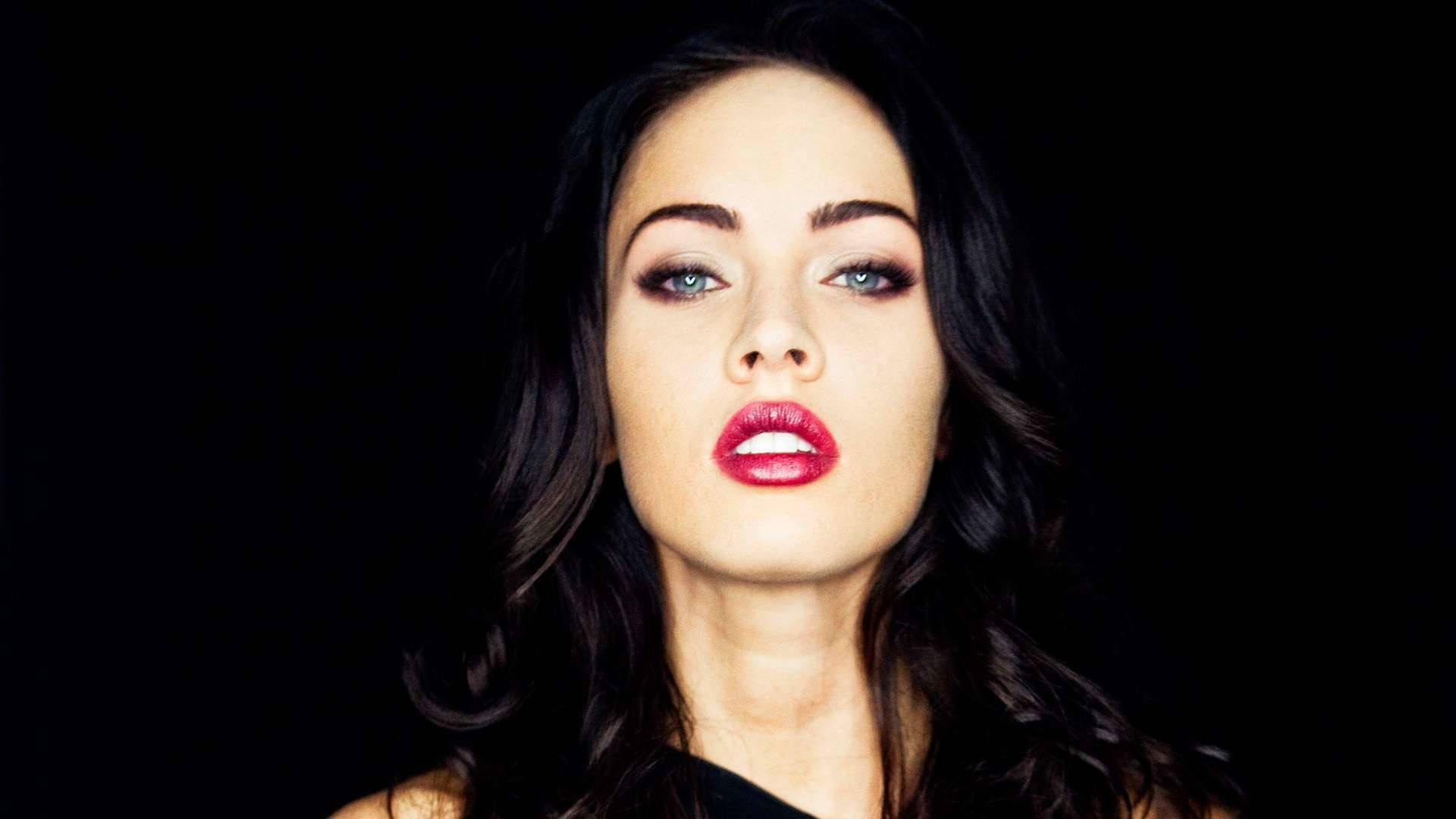 Megan Fox HD 桌布 壁紙 — 一個引人注目的肖像，一位女性擁有鮮豔的紅唇和強烈的藍眼睛，背景是純黑色。