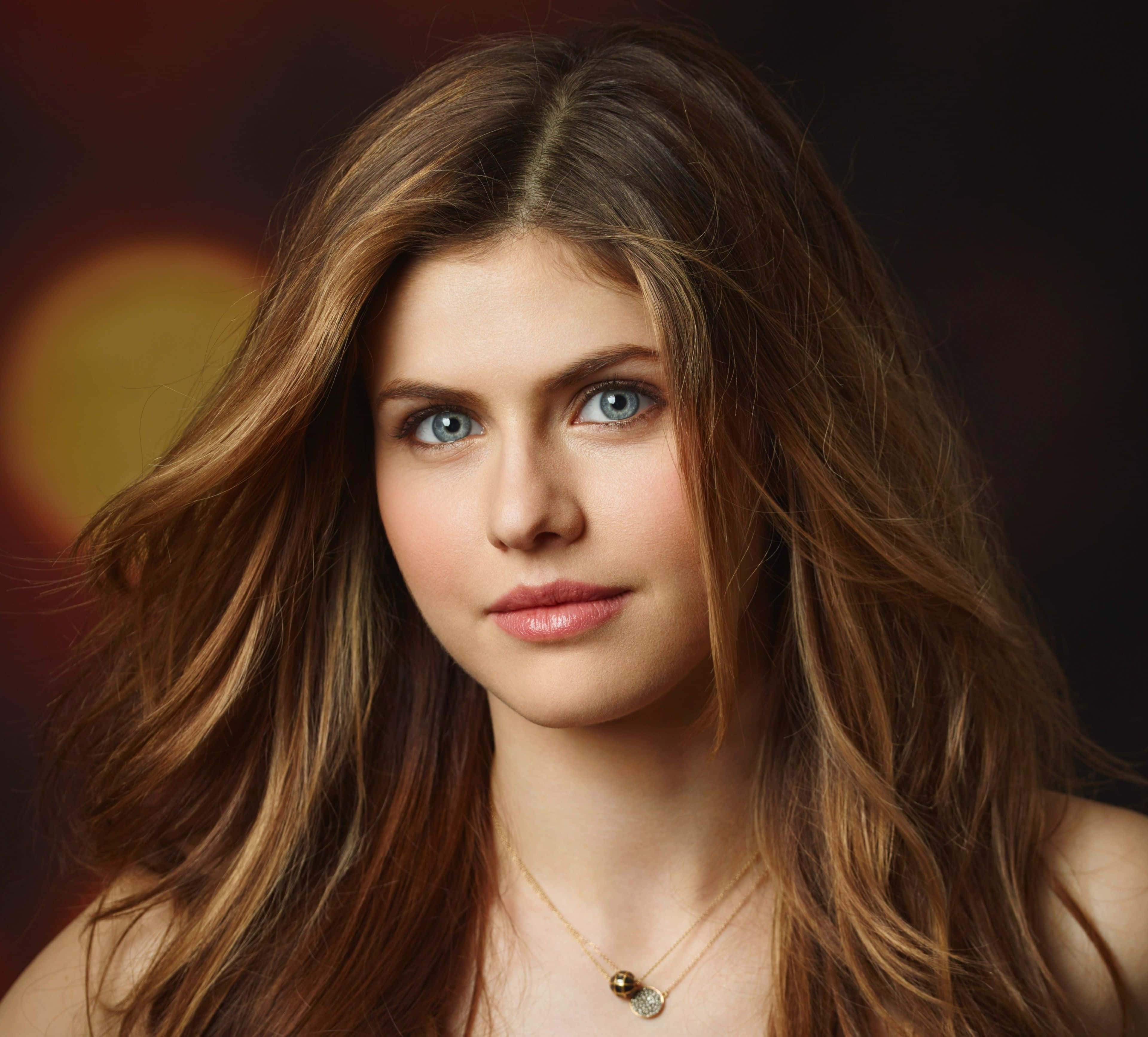 Alexandra Daddario 桌布 壁紙 — 一個引人注目的肖像，一位擁有鮮豔藍眼睛和飄逸棕色頭髮的女性，背景是溫暖模糊的。