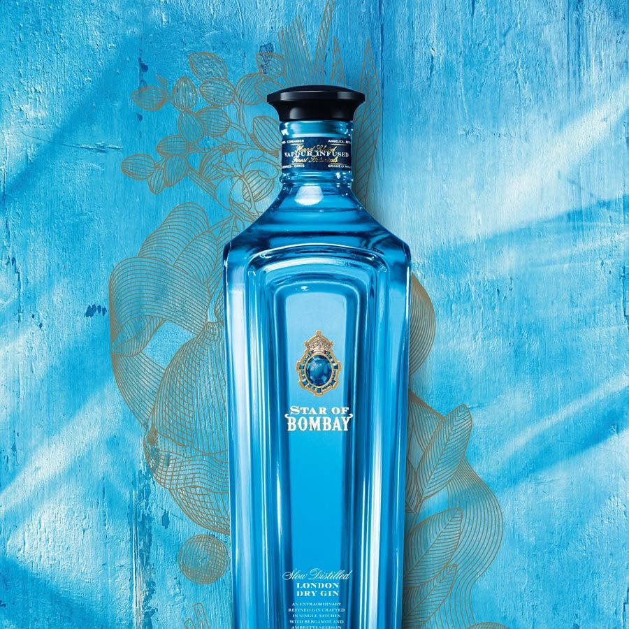 glass 桌布 壁紙 — 一個引人注目的藍色Star of Bombay琴酒瓶，靠在帶有金色植物蝕刻圖案的紋理藍色牆壁上。