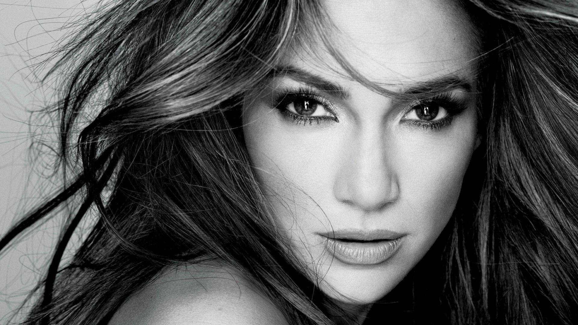 Jennifer Lopez 桌布 壁紙 — 一個引人注目的黑白特寫肖像，捕捉了一位頭髮隨風飄揚、眼神強烈的女性。