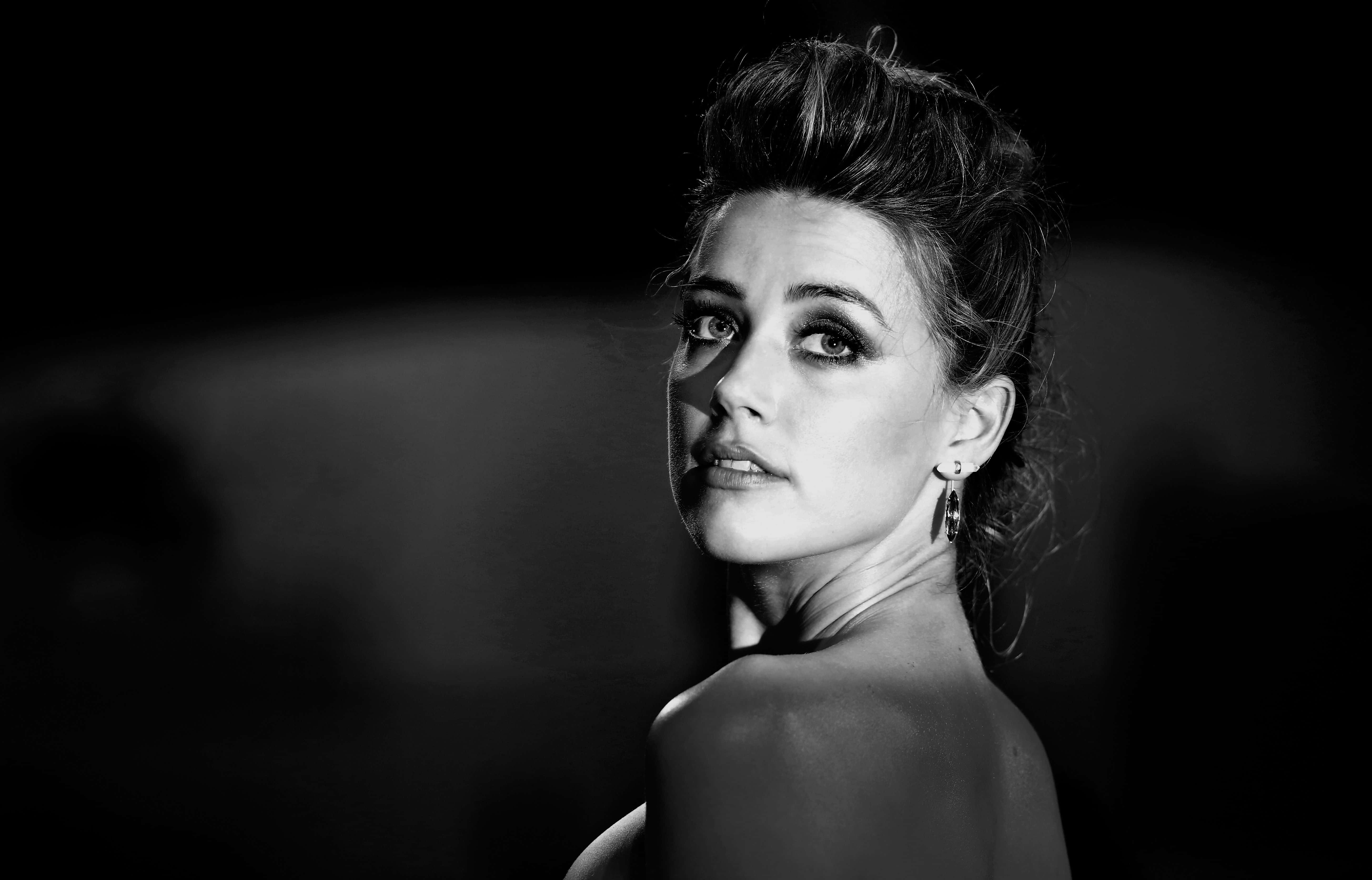 Amber Heard 桌布 壁紙 — 一個引人注目的黑白肖像，一位化著戲劇性妝容的女性在黑暗模糊的背景前回頭凝視。