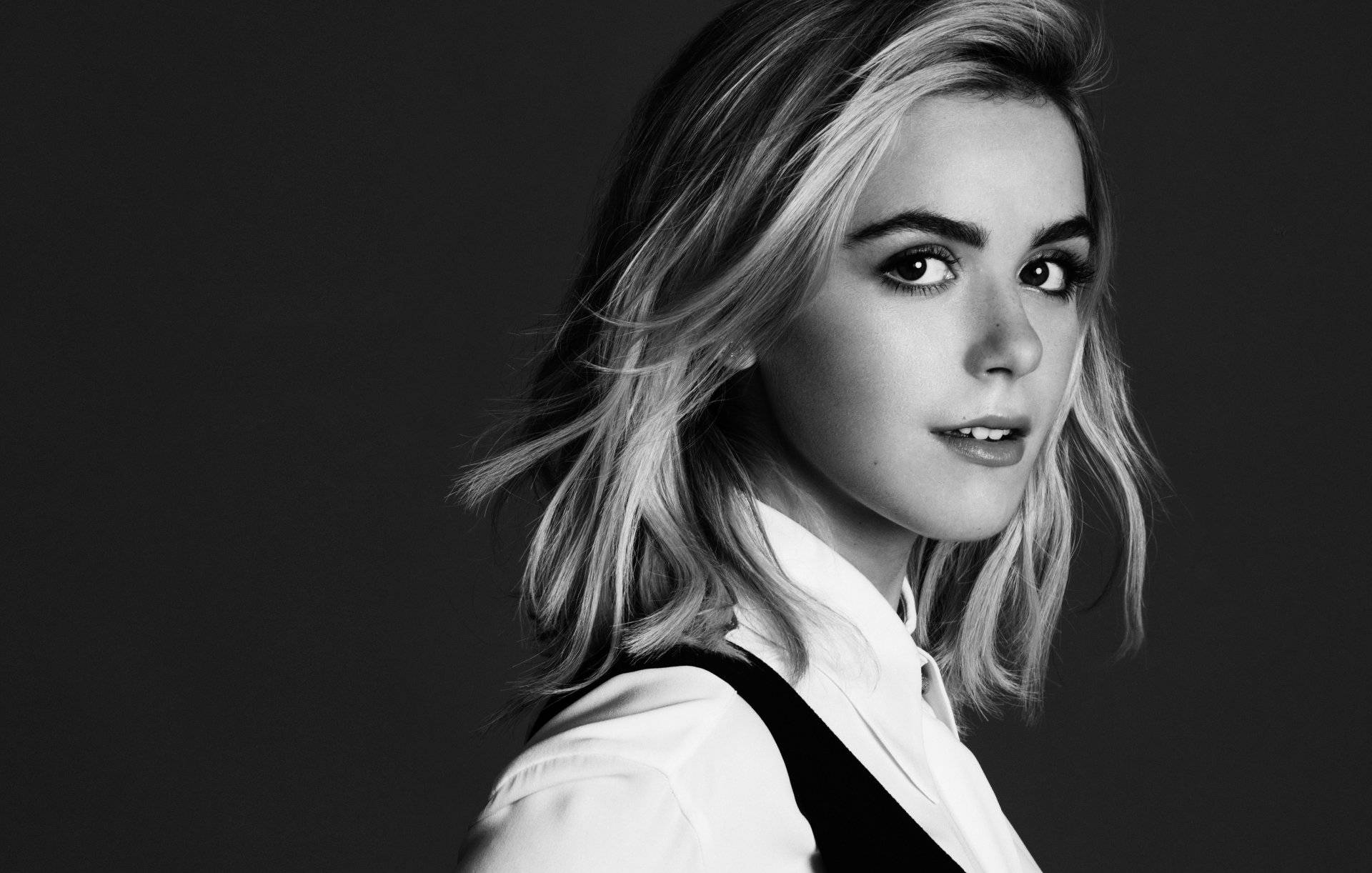 Kiernan Shipka 桌布 壁紙 — 一個引人注目的黑白肖像，一位年輕女性一頭凌亂的金髮，在黑暗背景下專注地凝視著相機。