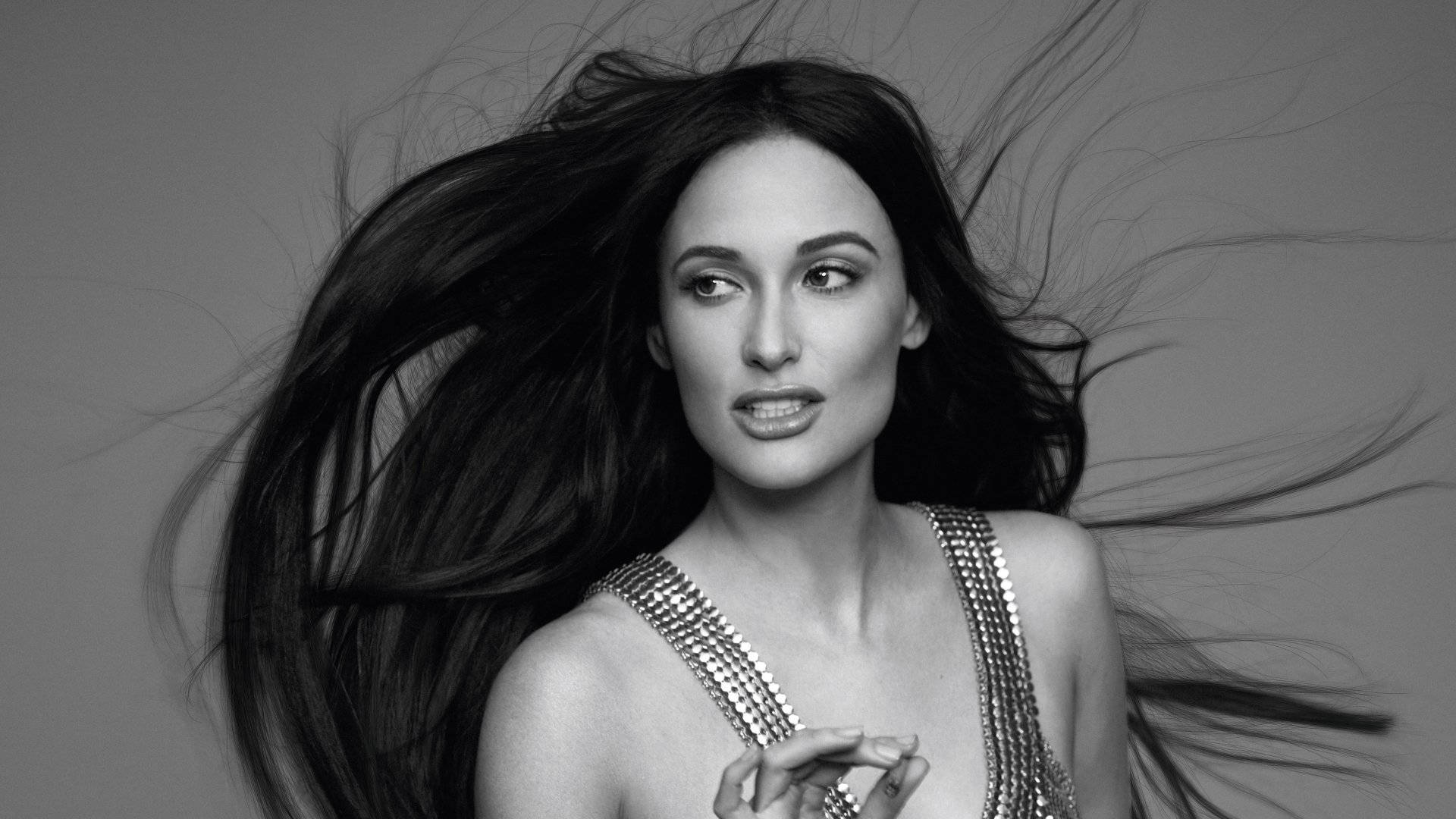 Kacey Musgraves 桌布 壁紙 — 一個引人注目的黑白肖像，一位擁有飄逸深色頭髮和閃亮上衣的女性，背景是純灰色。