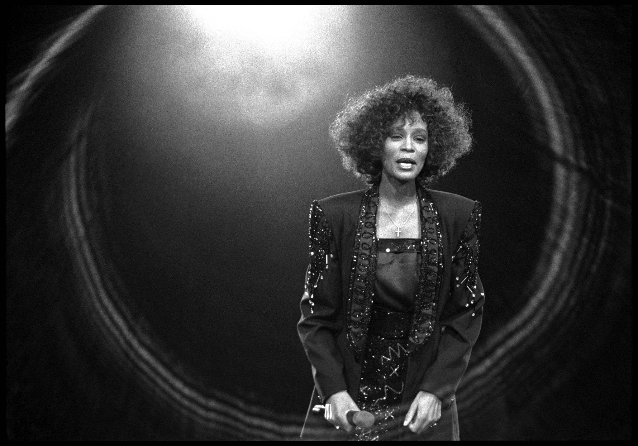 Whitney 桌布 壁紙 — 一個引人注目的黑白肖像，一位擁有豐盈捲髮的女性在聚光燈下唱歌，舞台上有同心圓形燈光效果。