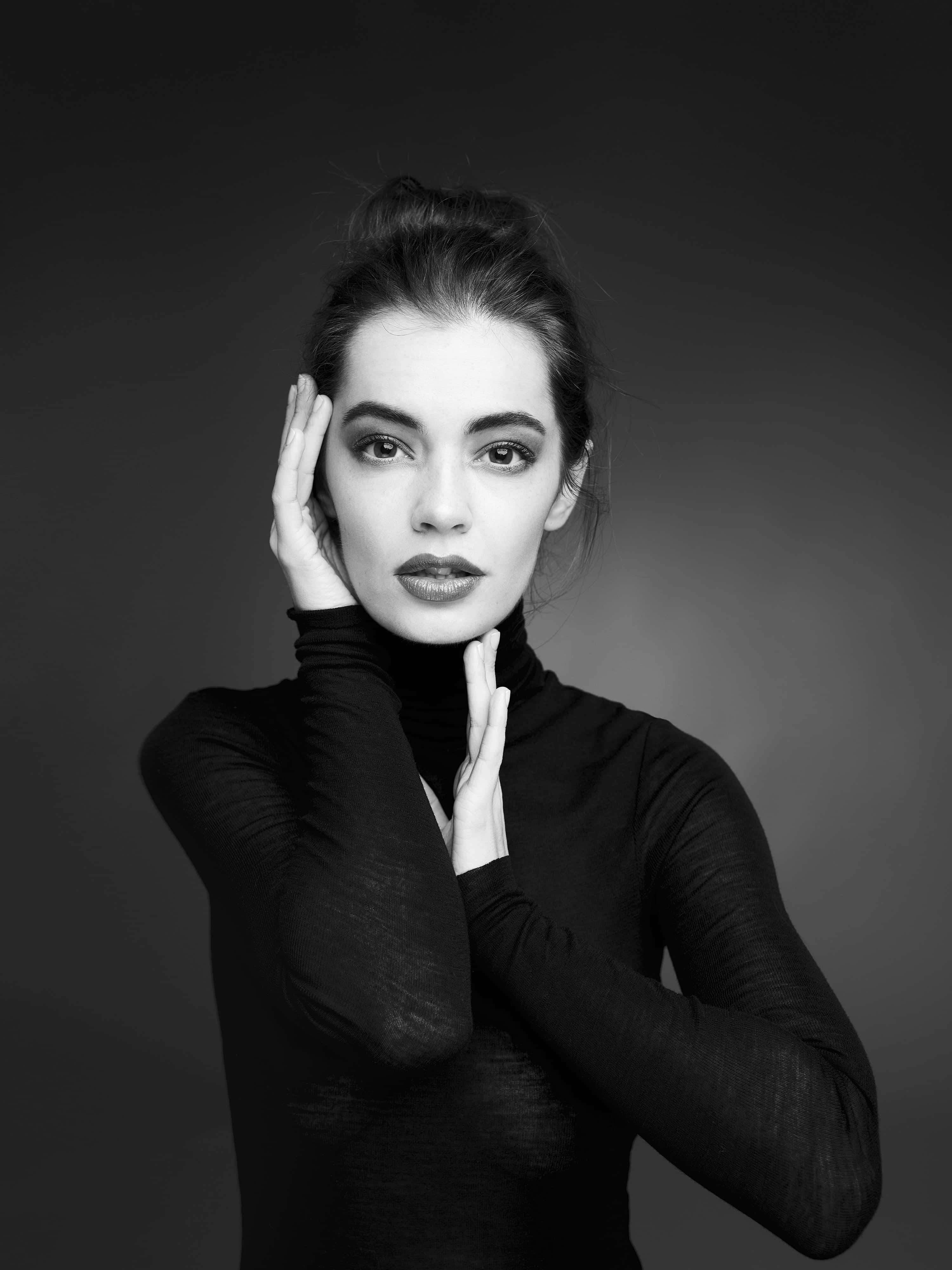 Black And White Fashion 桌布 壁紙 — 一個引人注目的黑白肖像，一位穿著高領毛衣的女性，雙手框住她的臉，背景是漸變灰色。