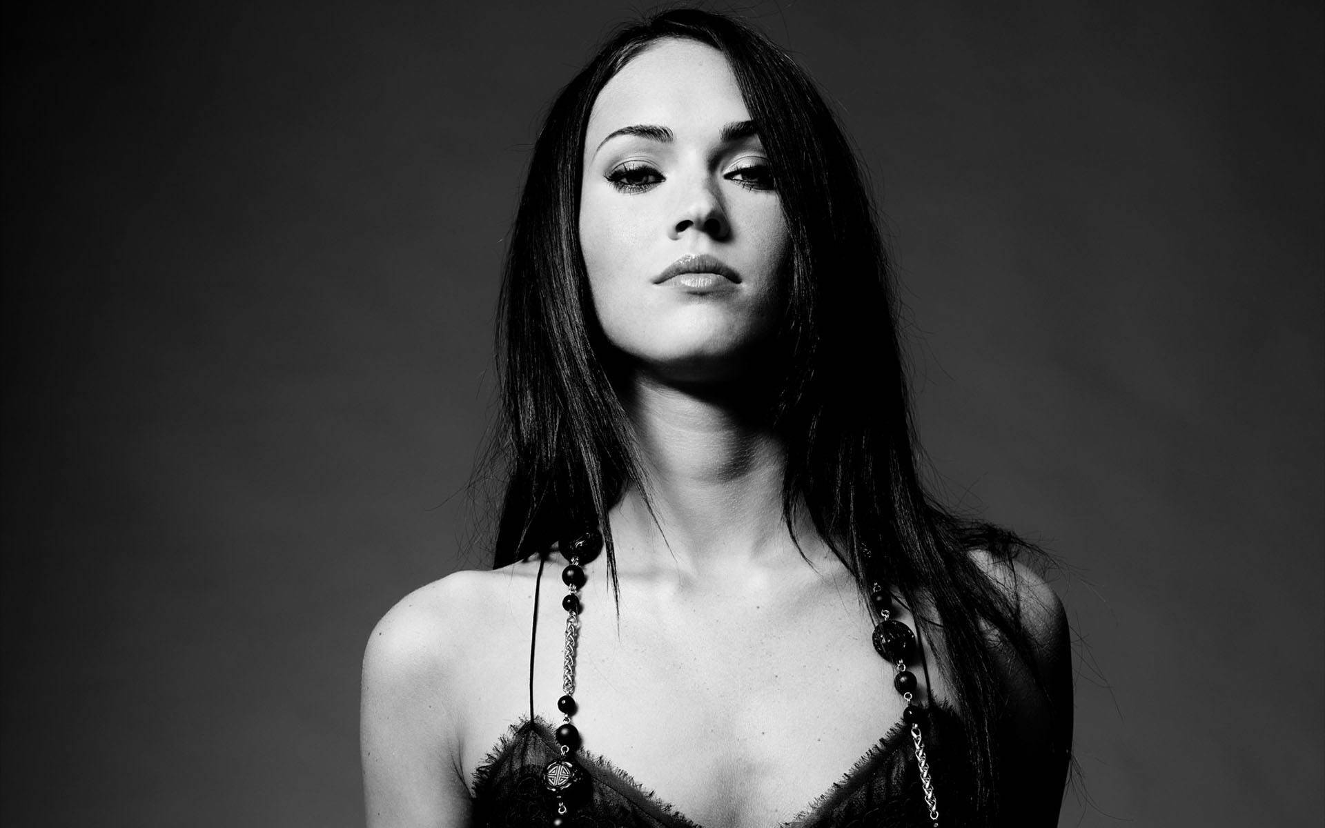 Megan Fox HD 桌布 壁紙 — 一個引人注目的黑白肖像，一位長黑髮的女性在純灰色背景下直視鏡頭。
