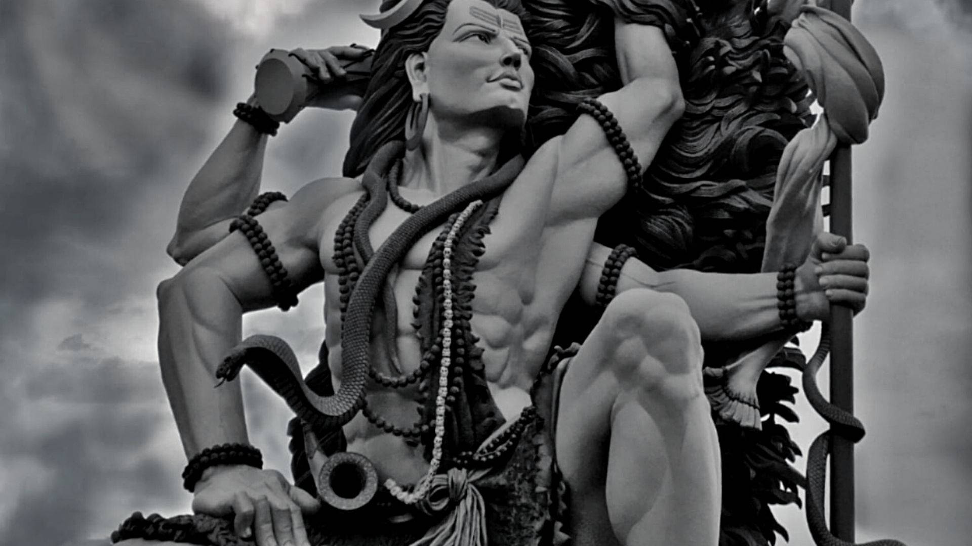 shiva dark 桌布 壁紙 — 一個強而有力的黑白雕像，描繪一位肌肉發達、長髮及帶有蛇形裝飾的神祇，背景是陰雲密布的天空。