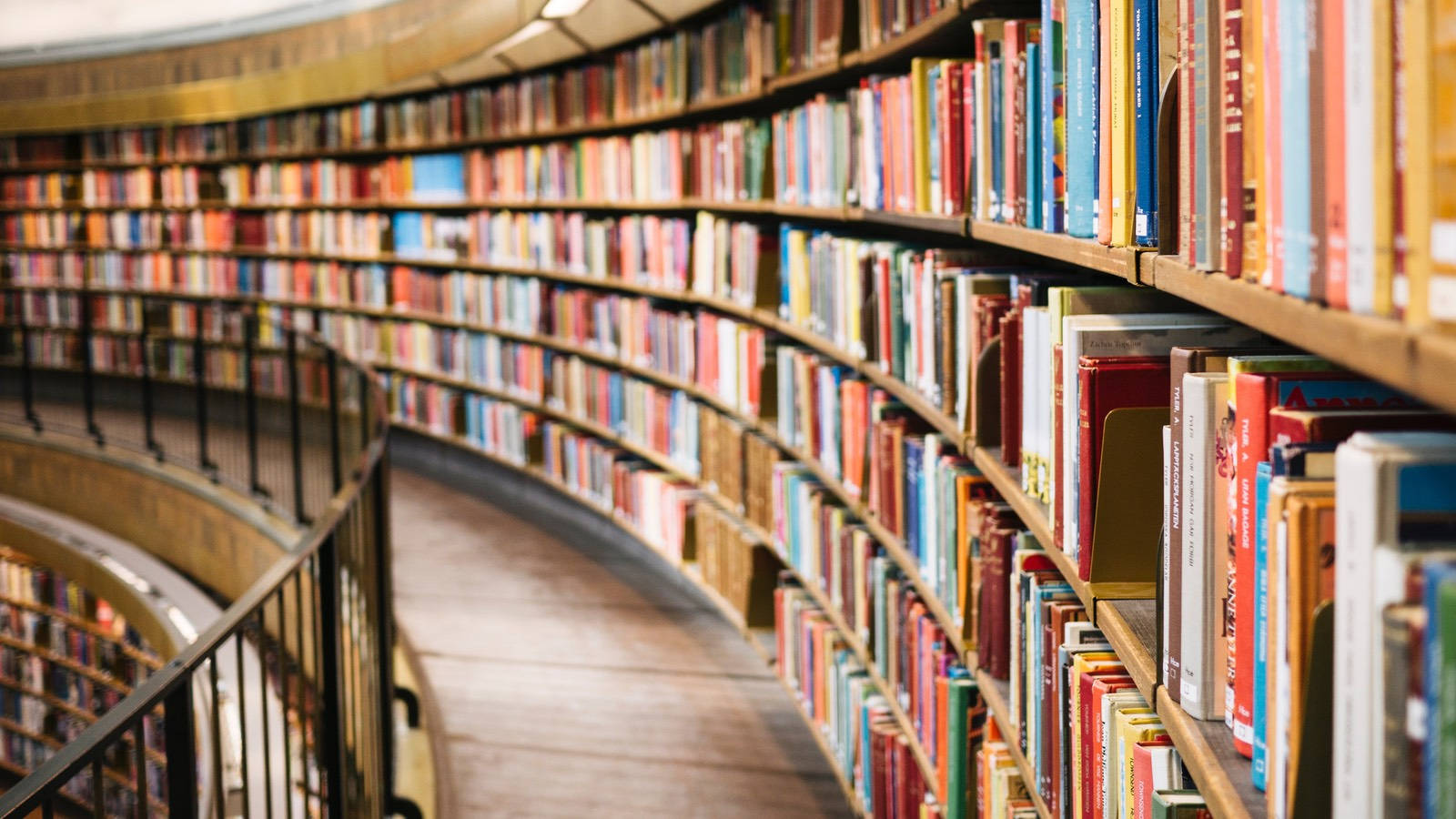 Library 桌布 壁紙 — 一個彎曲的木書架，裝滿色彩繽紛的書籍，在溫暖照明的圖書館通道中螺旋向上。