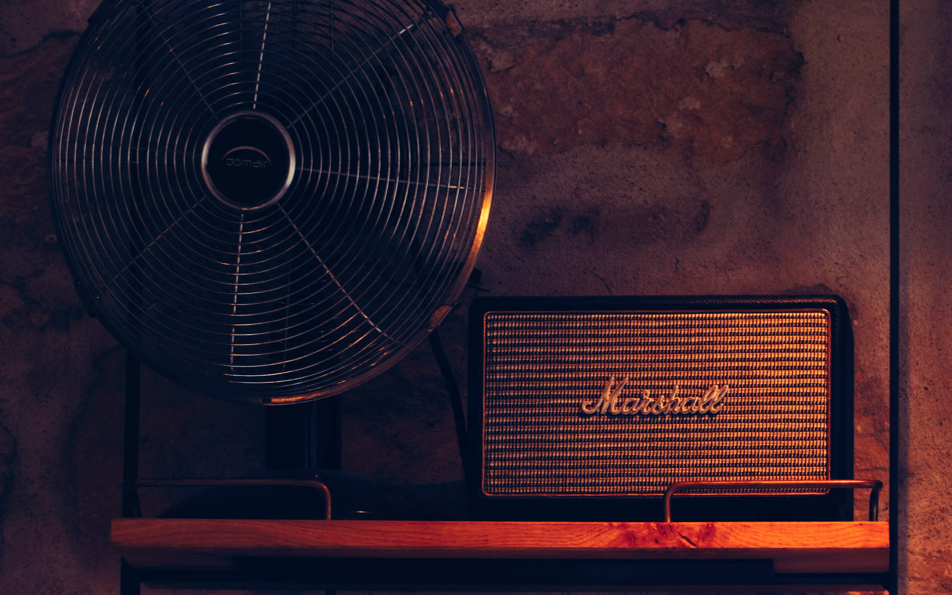 Marshall 桌布 壁紙 — 一個復古Marshall揚聲器和黑色風扇放在木架上，背景是紋理磚牆，在溫暖的環境光下。