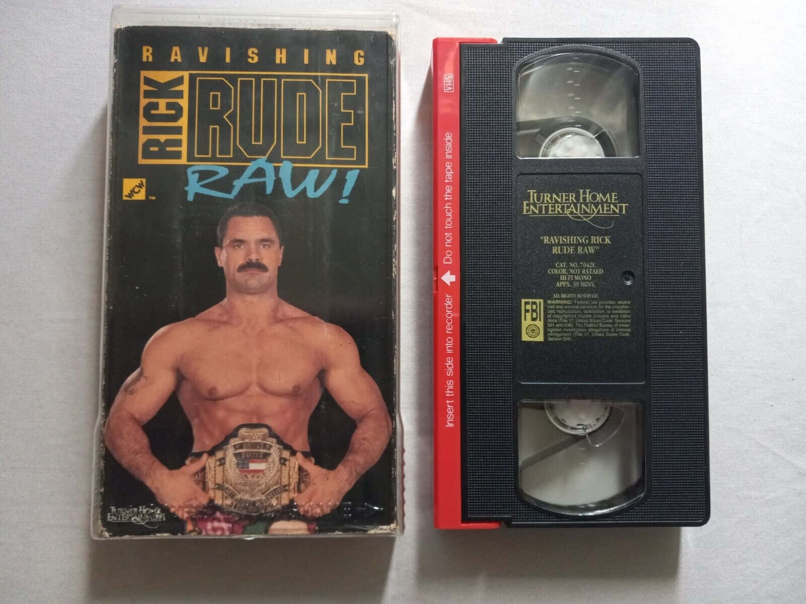 摔角 桌布 壁紙 — 一個復古VHS錄影帶及其盒子，用於「Ravishing Rick Rude Raw!」， featuring 摔角手在純白背景上持有冠軍腰帶。