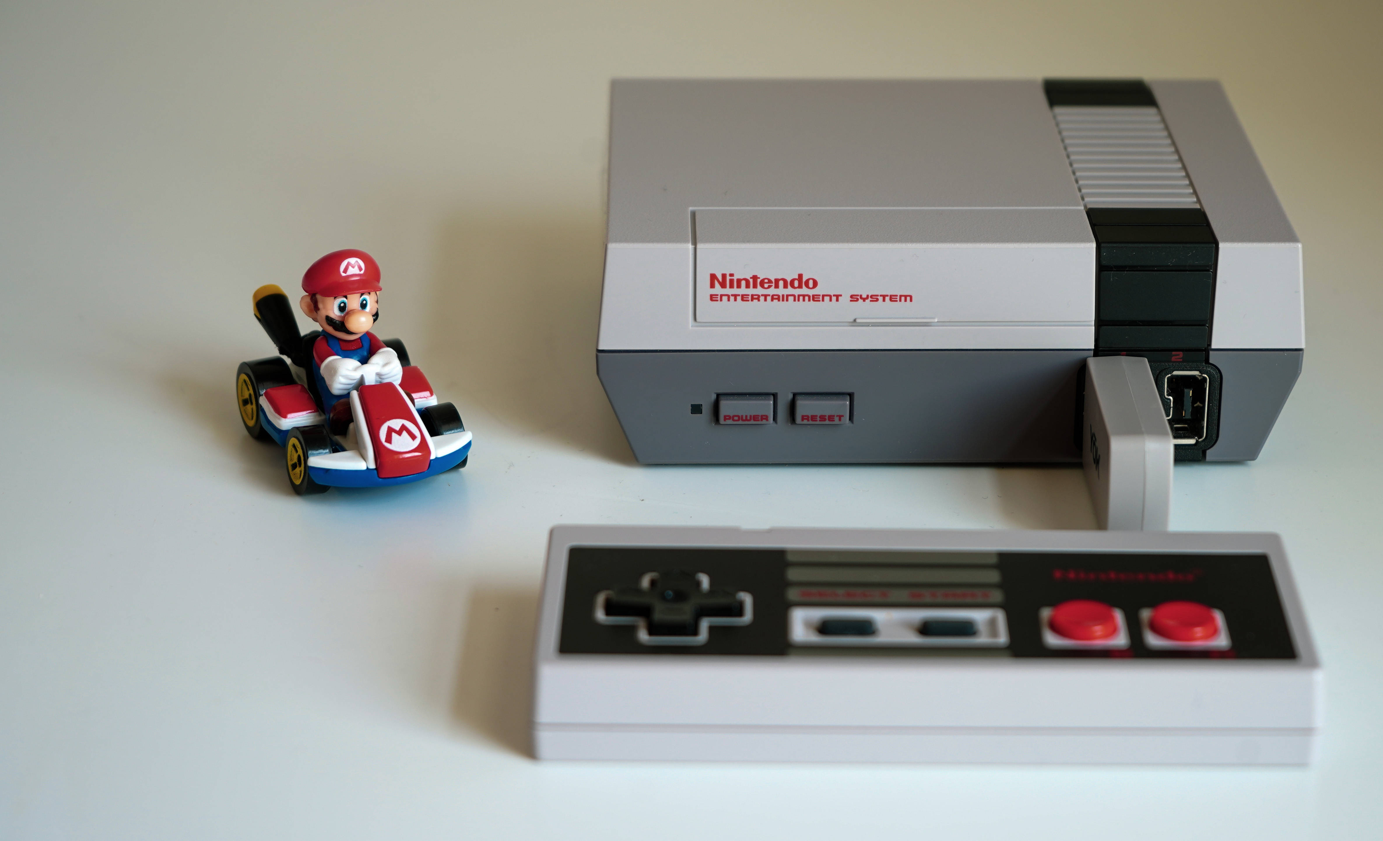 Nes 桌布 壁紙 — 一個復古任天堂娛樂系統主機，附有控制器和瑪利歐在卡丁車中的公仔，置於白色表面上。