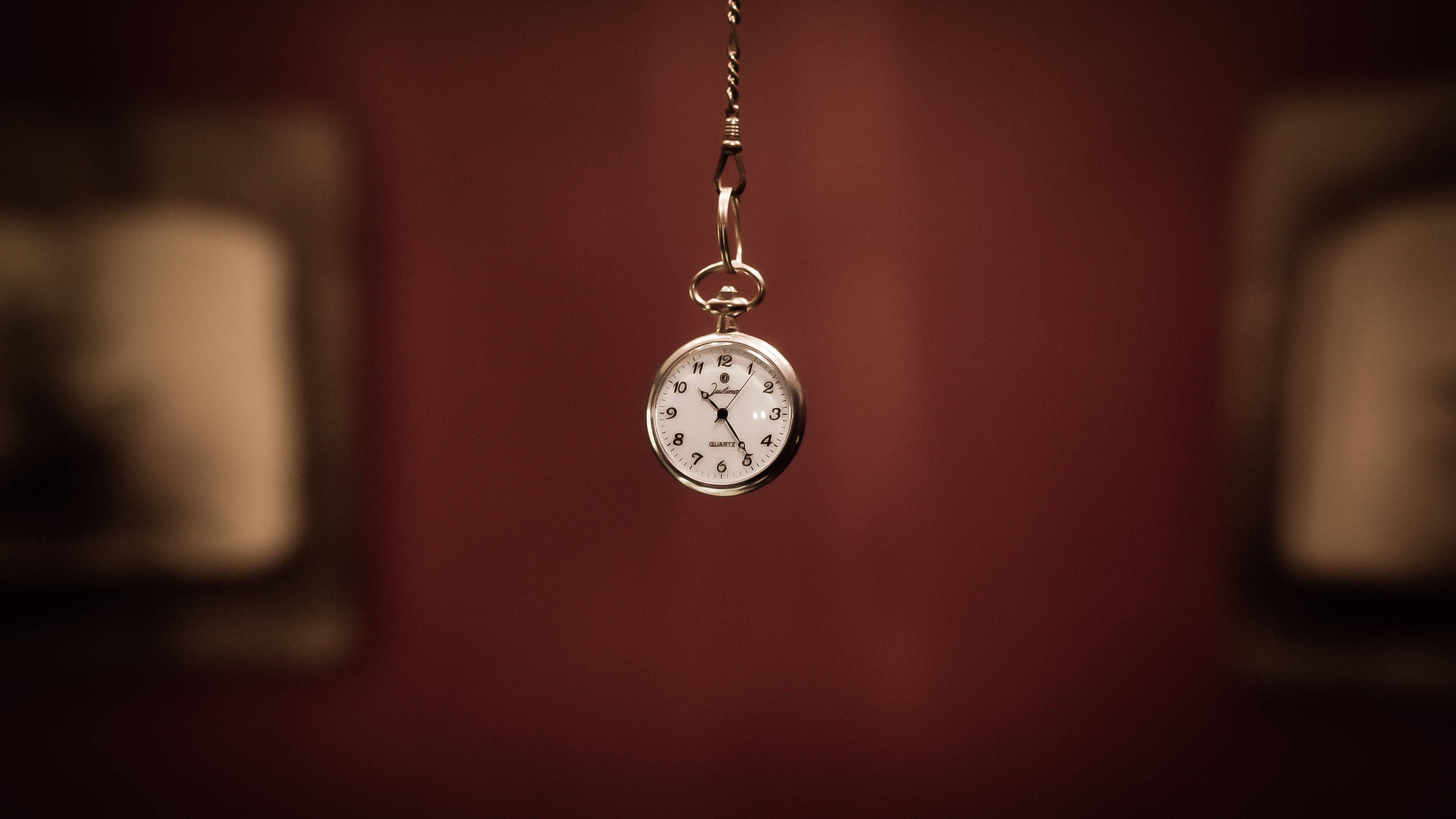 times up 桌布 壁紙 — 一個復古懷錶用鏈條懸掛在深紅色牆壁上，背景有模糊的相框圖片。