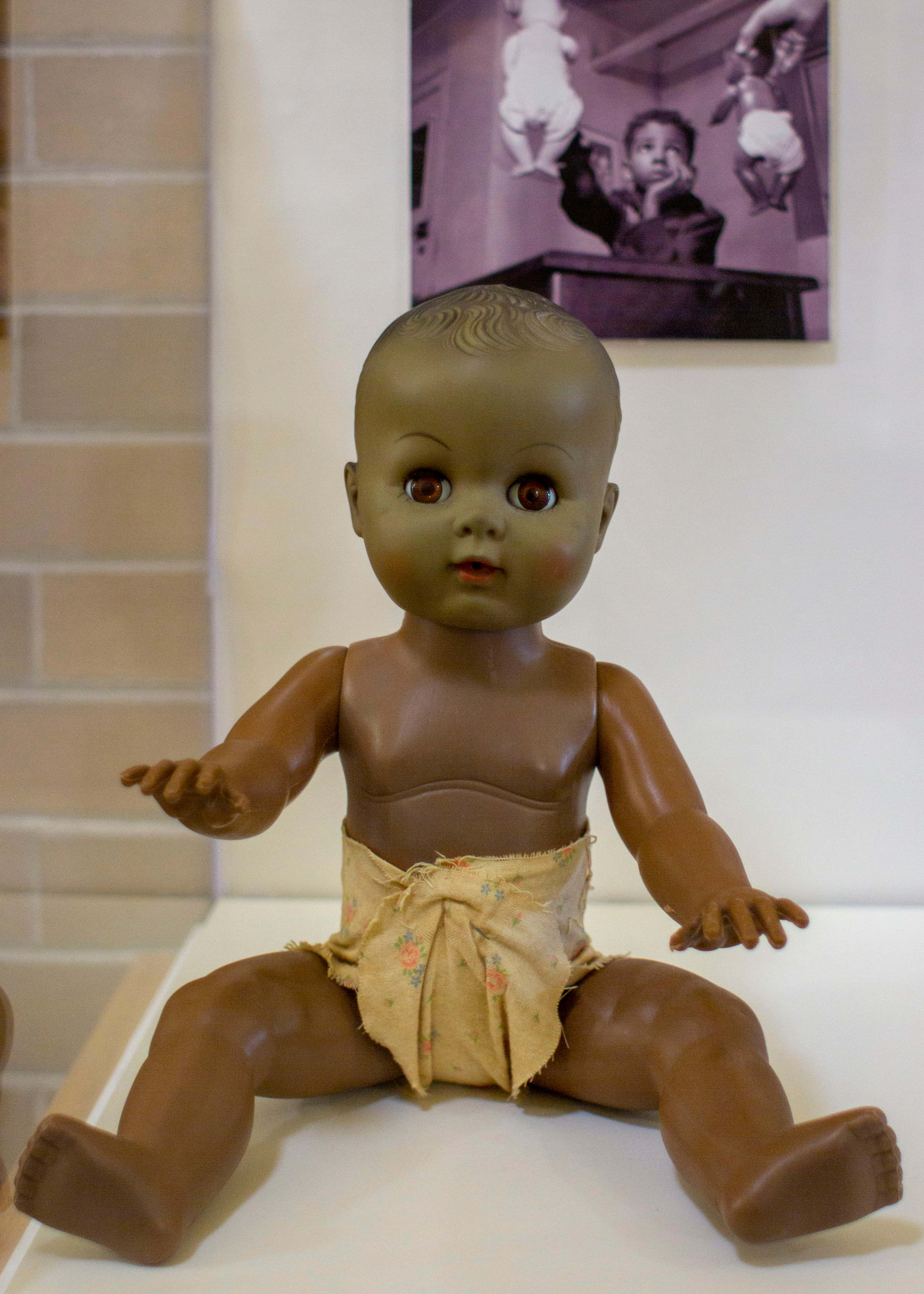 Doll 桌布 壁紙 — 一個復古棕色塑膠嬰兒娃娃，大眼睛，坐在白色表面上，穿著花卉尿布，背景有磚牆和裱框照片。