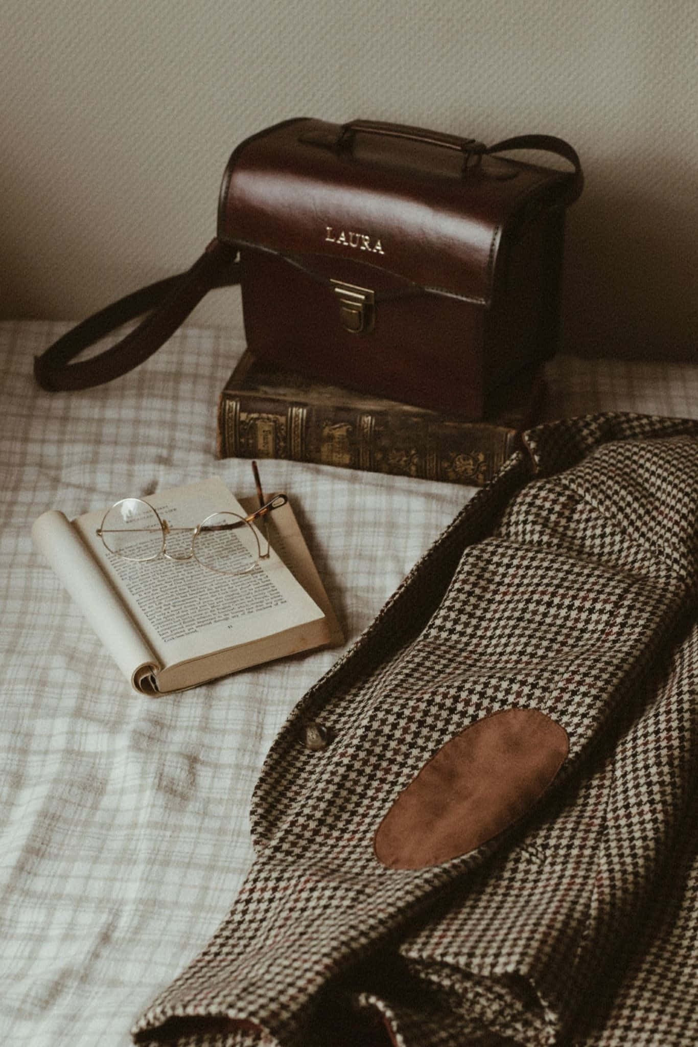 Dark Academia Aesthetic 桌布 壁紙 — 一個復古棕色皮革公事包，上面標有 laura，放在一本舊書上，旁邊有一本打開的書和眼鏡，放在格子床單上。