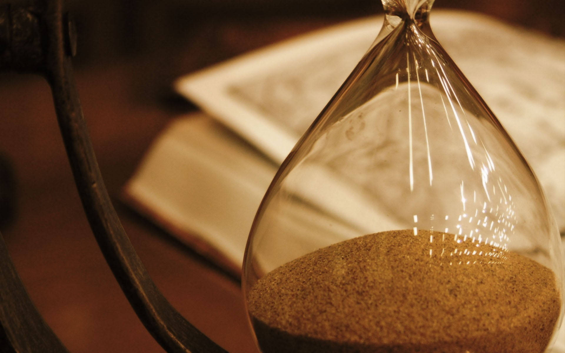 Hourglass 桌布 壁紙 — 一個復古沙漏，金色沙子緩緩流下，旁邊放著一本打開的書，光線溫暖柔和。