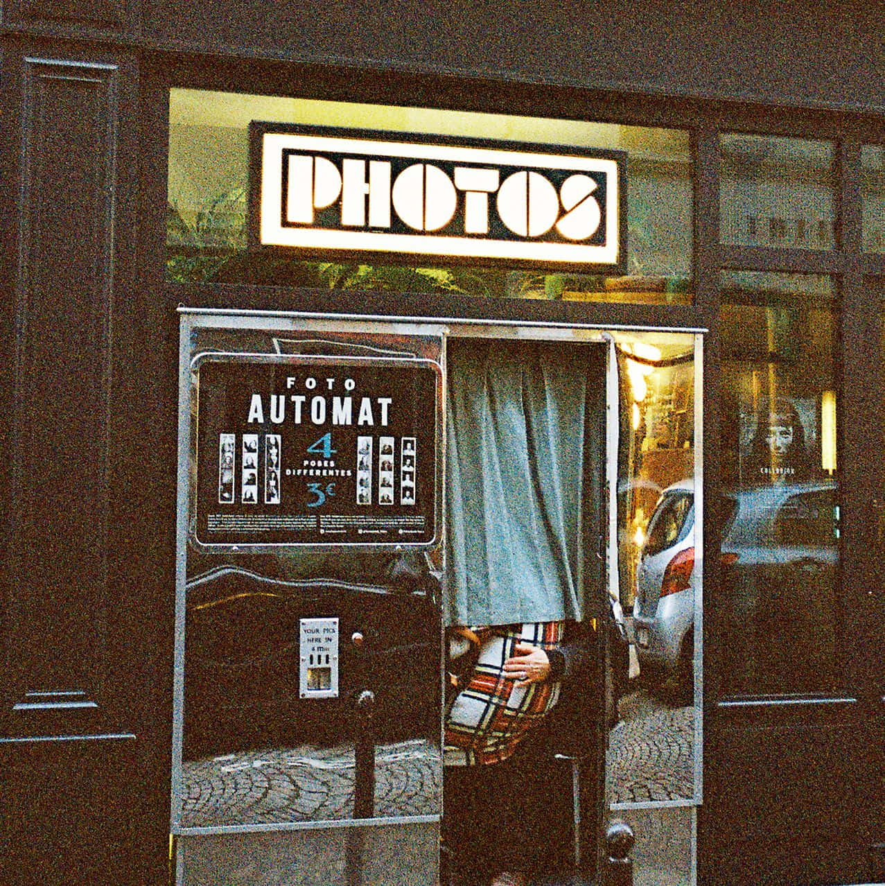 Photo Booth 桌布 壁紙 — 一個復古照相亭，帶有發光的「PHOTOS」標誌，裡面有一個穿著格子衣服的人拿著相機。