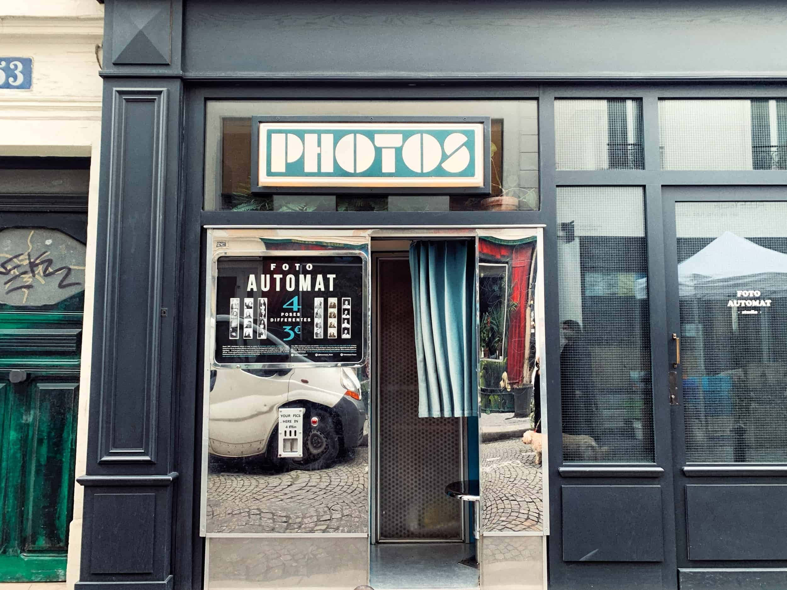 Photo Booth 桌布 壁紙 — 一個復古照相亭，帶有青綠色招牌和藍色窗簾，矗立在鵝卵石街道上，反射出一輛停著的汽車和城市環境。
