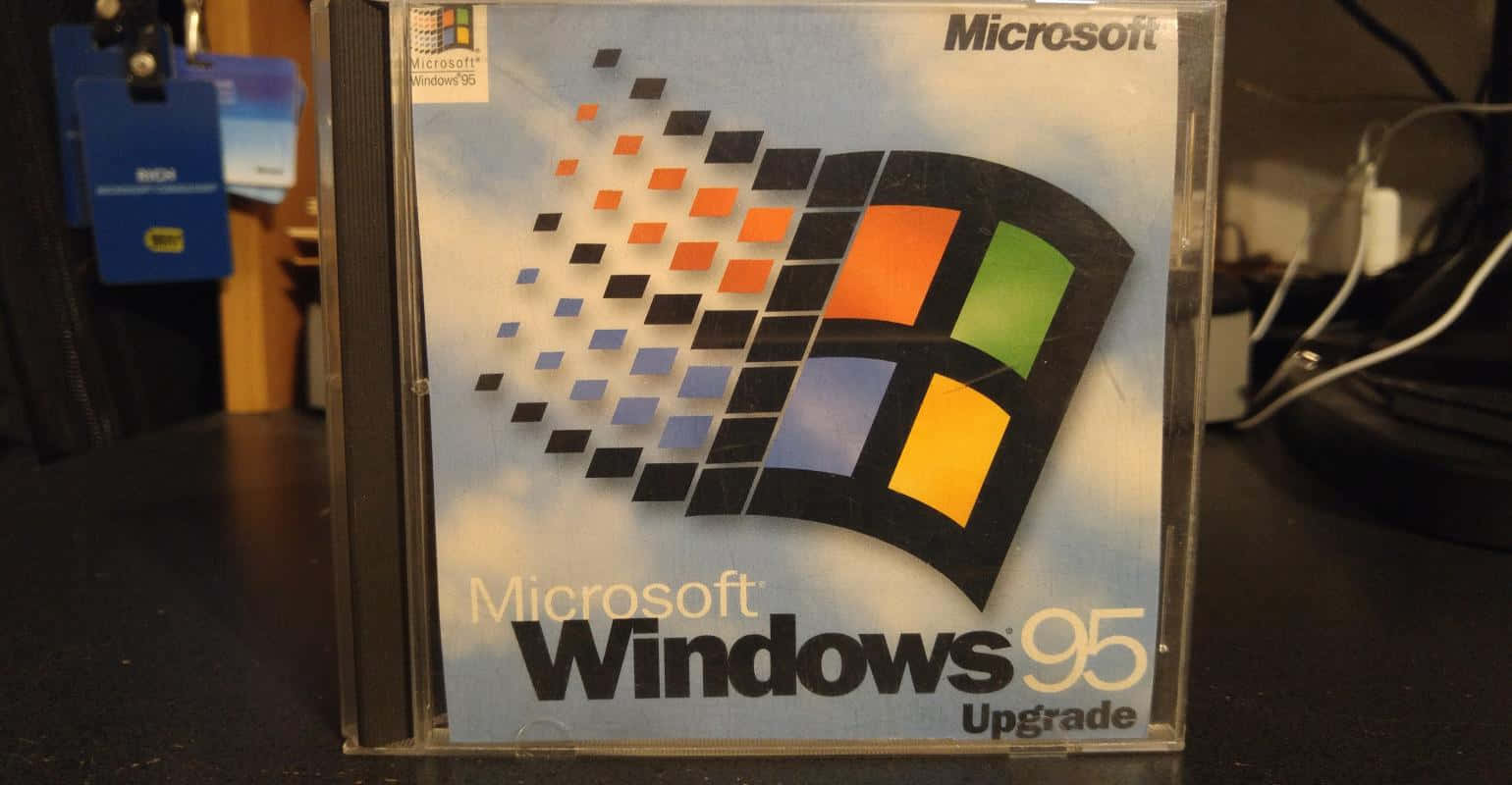 Windows 95 動態圖 桌布 壁紙 — 一個復古的 Microsoft Windows 95 升級版 CD 盒放在深色桌子上，背景是模糊的科技背景。
