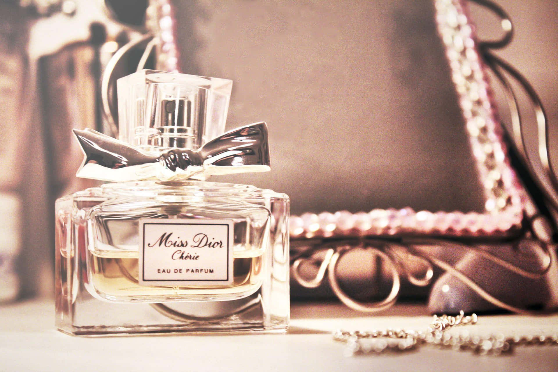 Dior 動態 桌布 壁紙 — 一個復古的 Miss Dior Chérie 香水瓶，帶有銀色蝴蝶結，優雅地放在一個柔光照射的表面上，旁邊是一個珠飾手袋和珠寶。