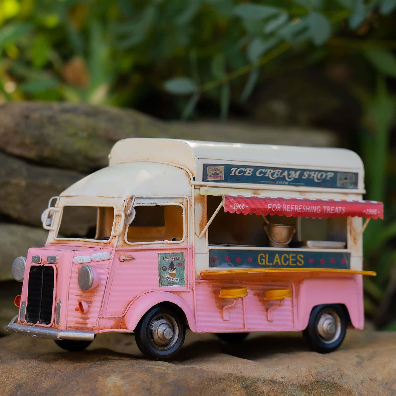 Ice Cream Truck 桌布 壁紙 — 一個復古粉紅色和白色的冰淇淋車模型放在岩石上，背景有柔焦的綠色樹葉。