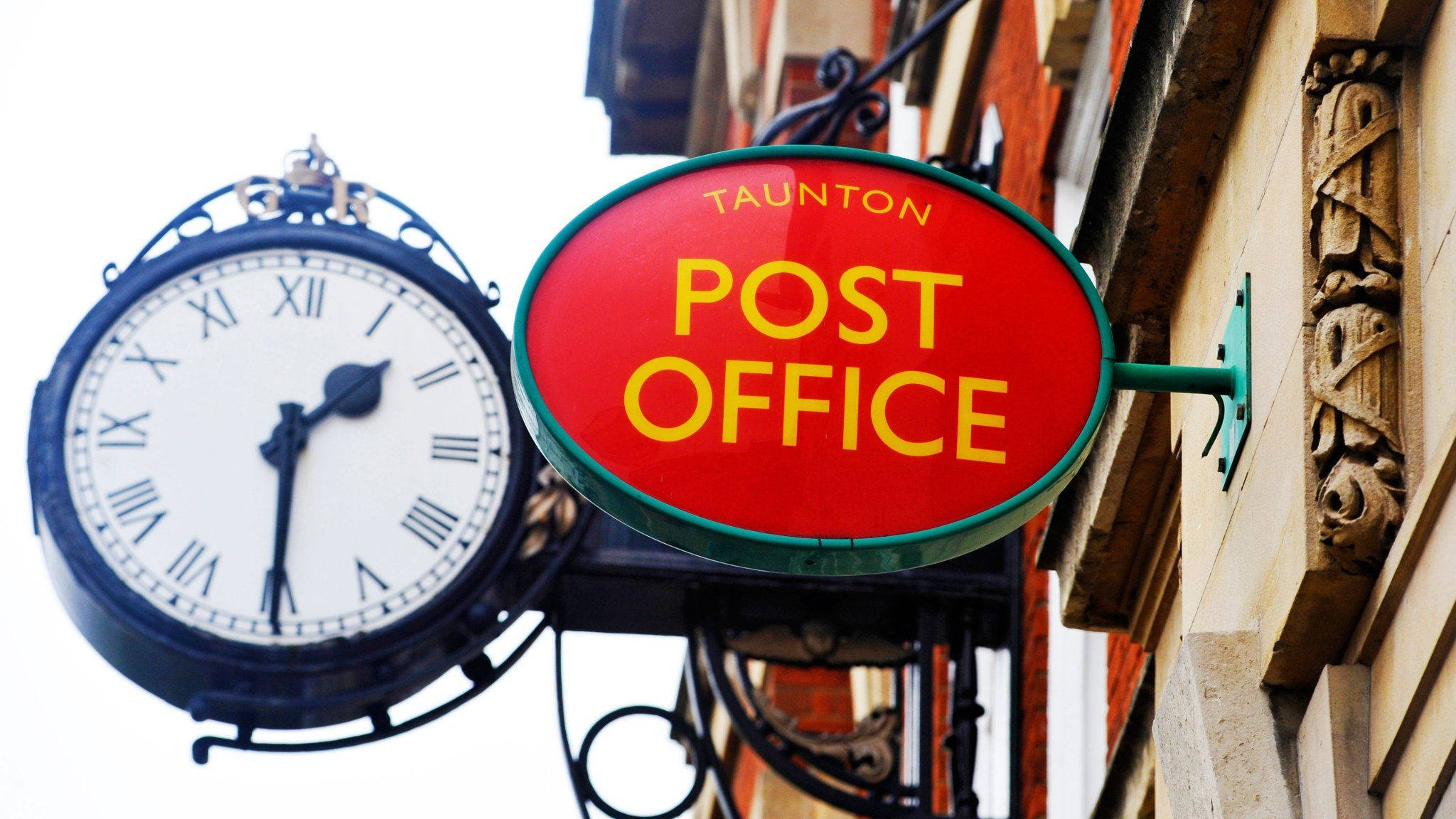 Post Office 桌布 壁紙 — 一個復古紅色橢圓形郵政局標誌，帶有黃色字體，懸掛在歷史建築外牆上一個帶有羅馬數字的黑色華麗時鐘旁邊。