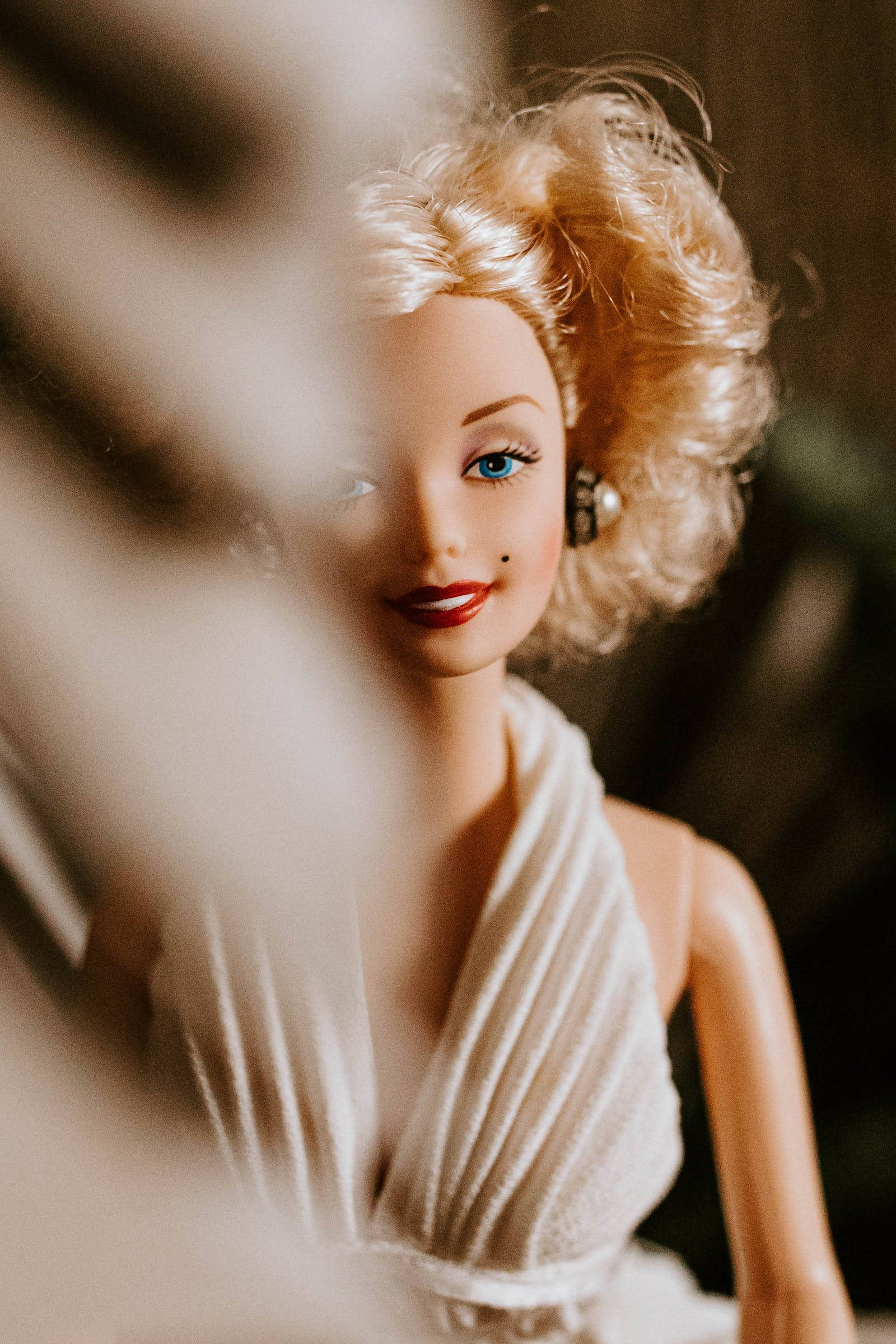Doll 桌布 壁紙 — 一個復古芭比娃娃，金色捲髮和紅唇，透過模糊的前景元素溫柔地凝視。